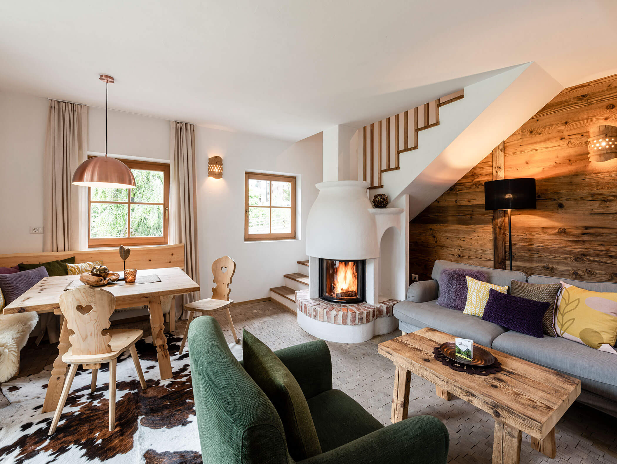 Gemütliches Wohnzimmer mit Kamin, Holzmöbeln, grünen Stühlen und einer Essecke unter einer Kupferlampe. - Maurlechnhof