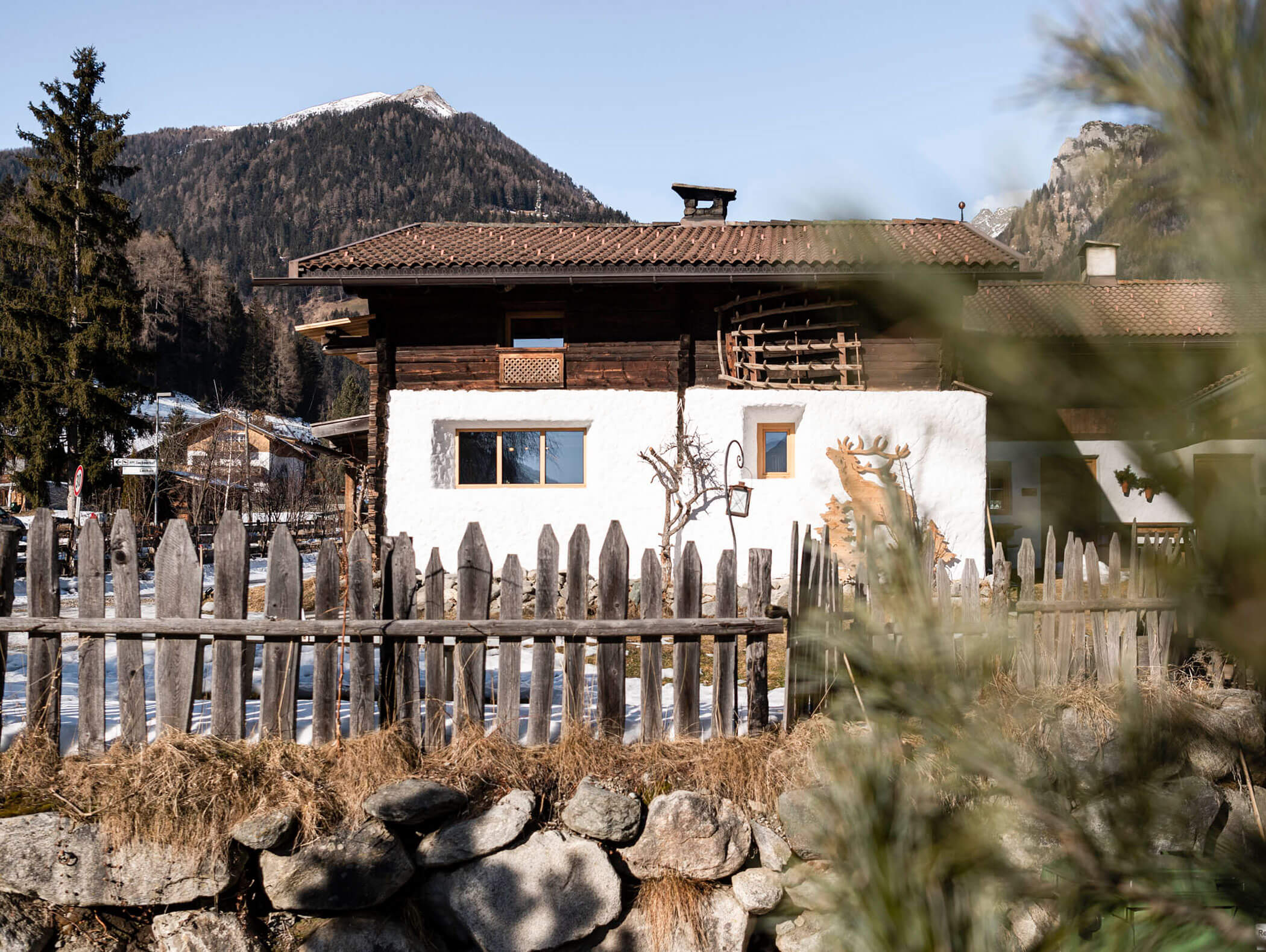 Ein rustikales Haus mit Holzzaun und Bergkulisse, umgeben von Bäumen und Felsen an einem sonnigen Tag. - Maurlechnhof