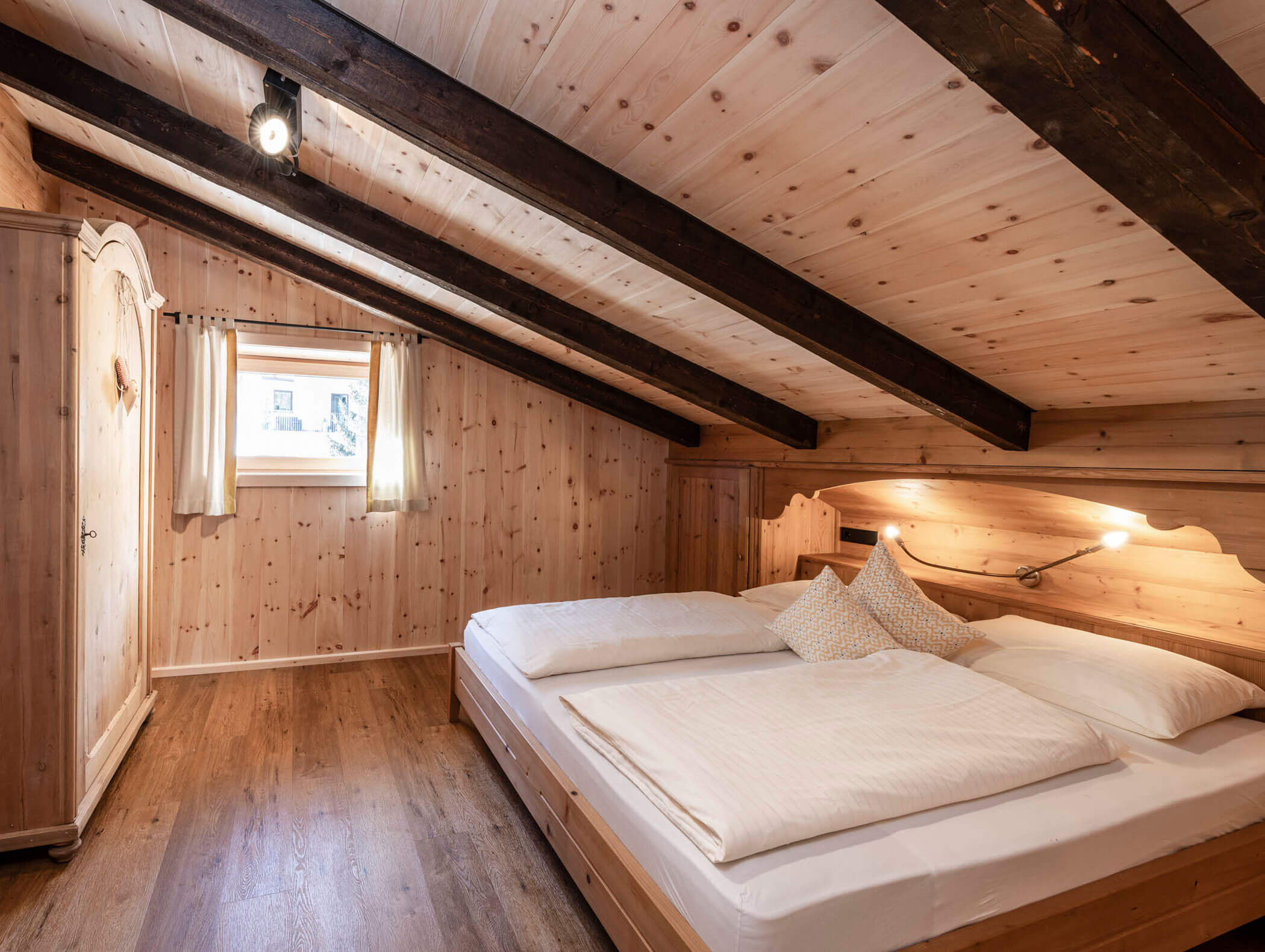 Gemütliches Schlafzimmer im Dachgeschoss mit Holzwänden, sichtbaren Balken, einem Doppelbett und einem kleinen Fenster mit Vorhängen. - Maurlechnhof