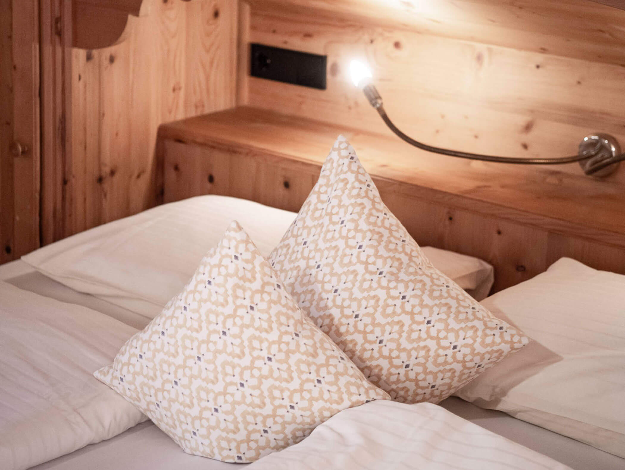 Zwei gemusterte Kissen auf einem ordentlich gemachten Bett mit weißer Bettwäsche und einem Holzkopfteil mit Leselicht. - Maurlechnhof