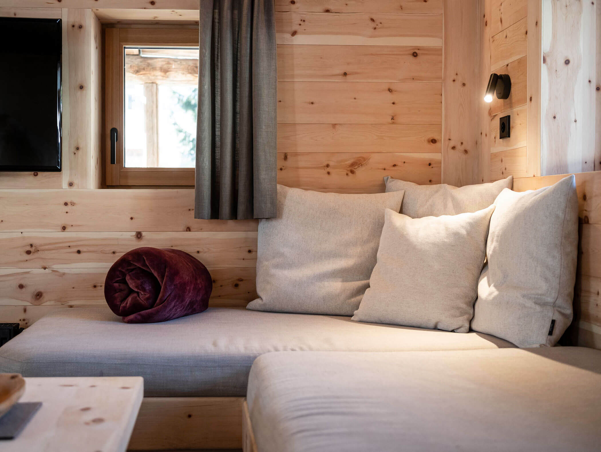 Gemütliches Holzzimmer mit einem gepolsterten Sofa, Wurfkissen, einer zusammengerollten Decke, einem Fenster und einer Wandlampe. - Maurlechnhof