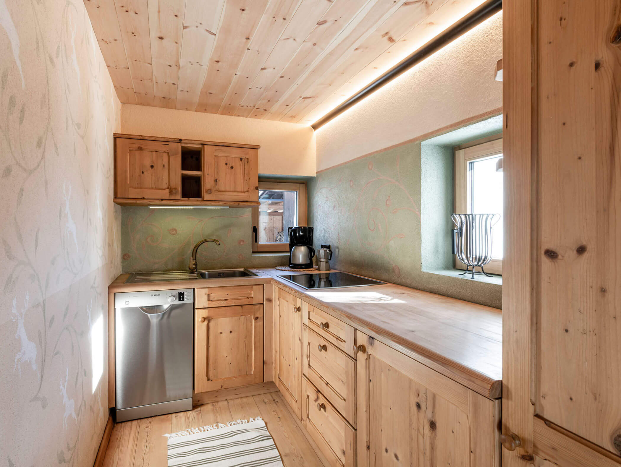 Kleine rustikale Küche mit Holzschränken, Geschirrspüler, Kaffeemaschine, Spülbecken und Fenster mit Tageslicht. - Maurlechnhof