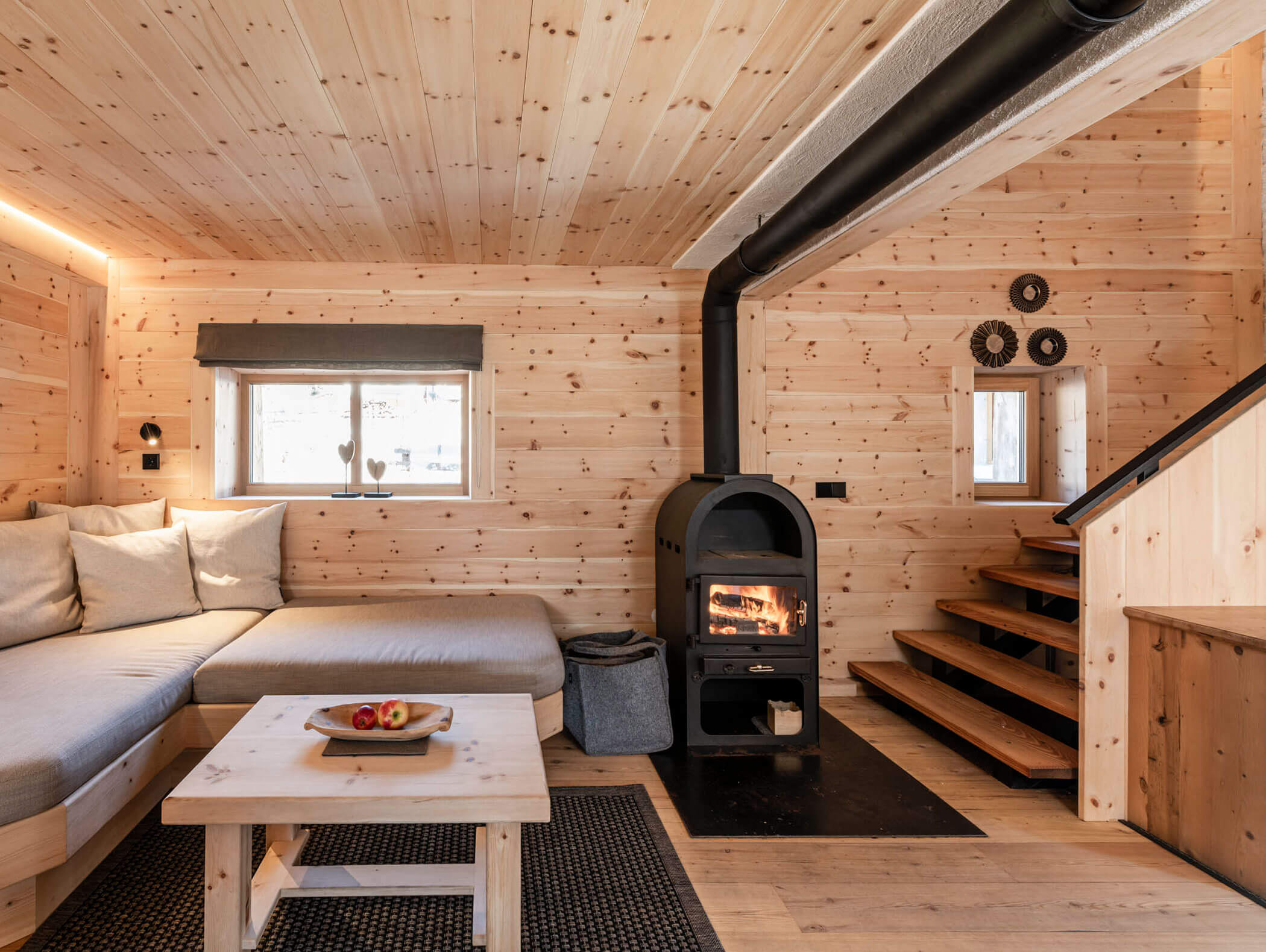 Gemütliche Holzhütte mit beleuchtetem Ofen, Ecksofa, Couchtisch und einer Treppe, die nach oben führt. - Maurlechnhof