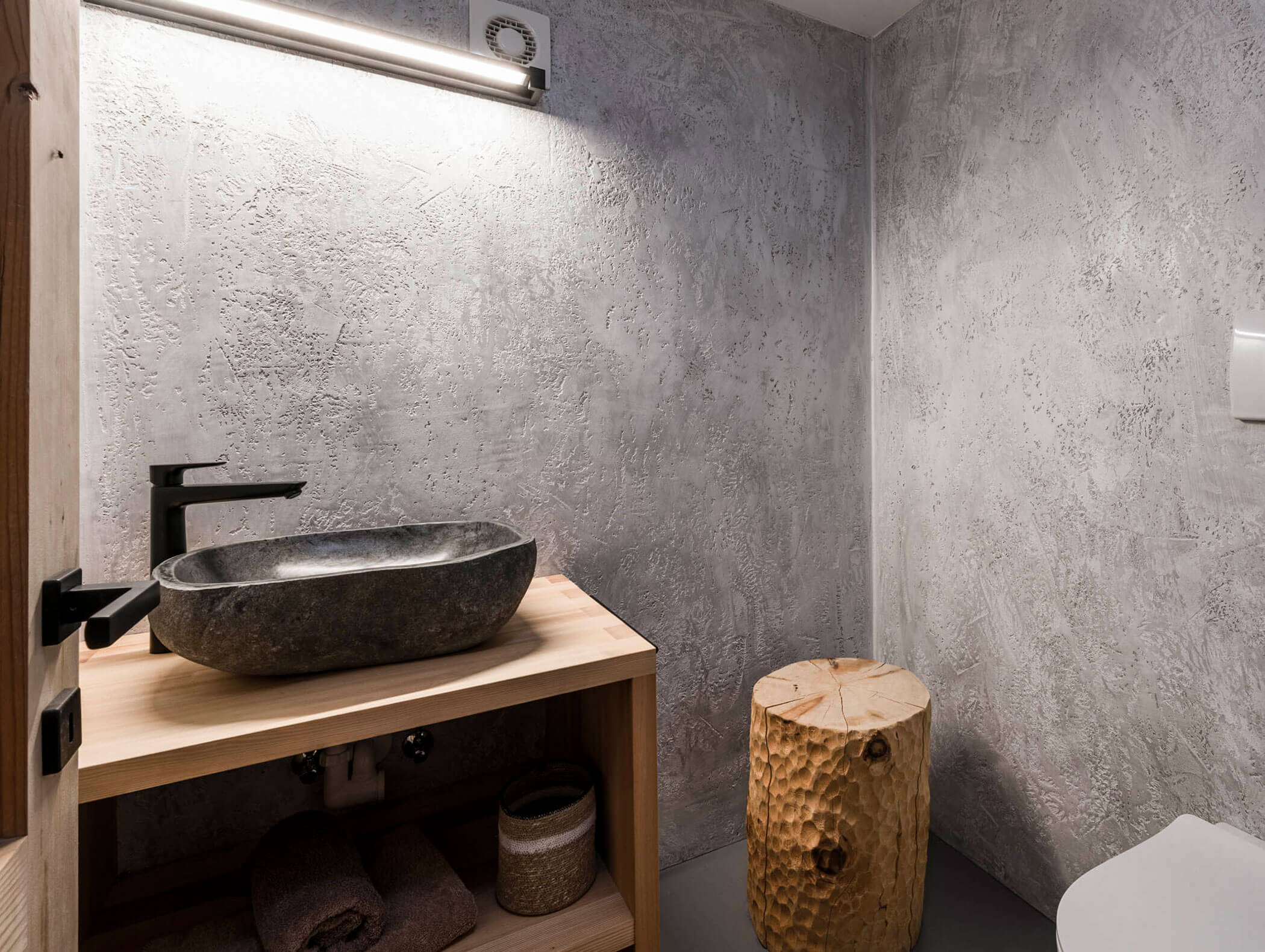 Modernes Badezimmer mit Steinwaschbecken, hölzernem Waschtisch, strukturierten Wänden und einem geschnitzten Holzhocker. - Maurlechnhof