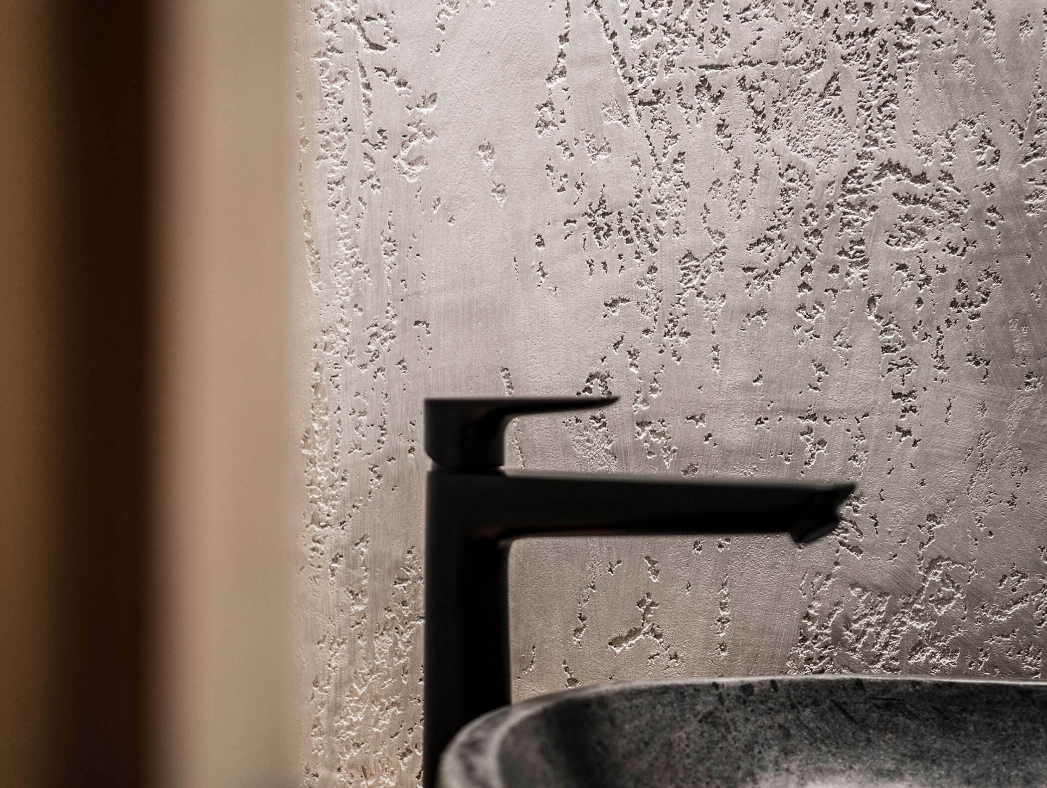 Schwarzer moderner Wasserhahn und Steinwaschbecken vor einer strukturierten beigen Wand mit rauem Putz. - Maurlechnhof