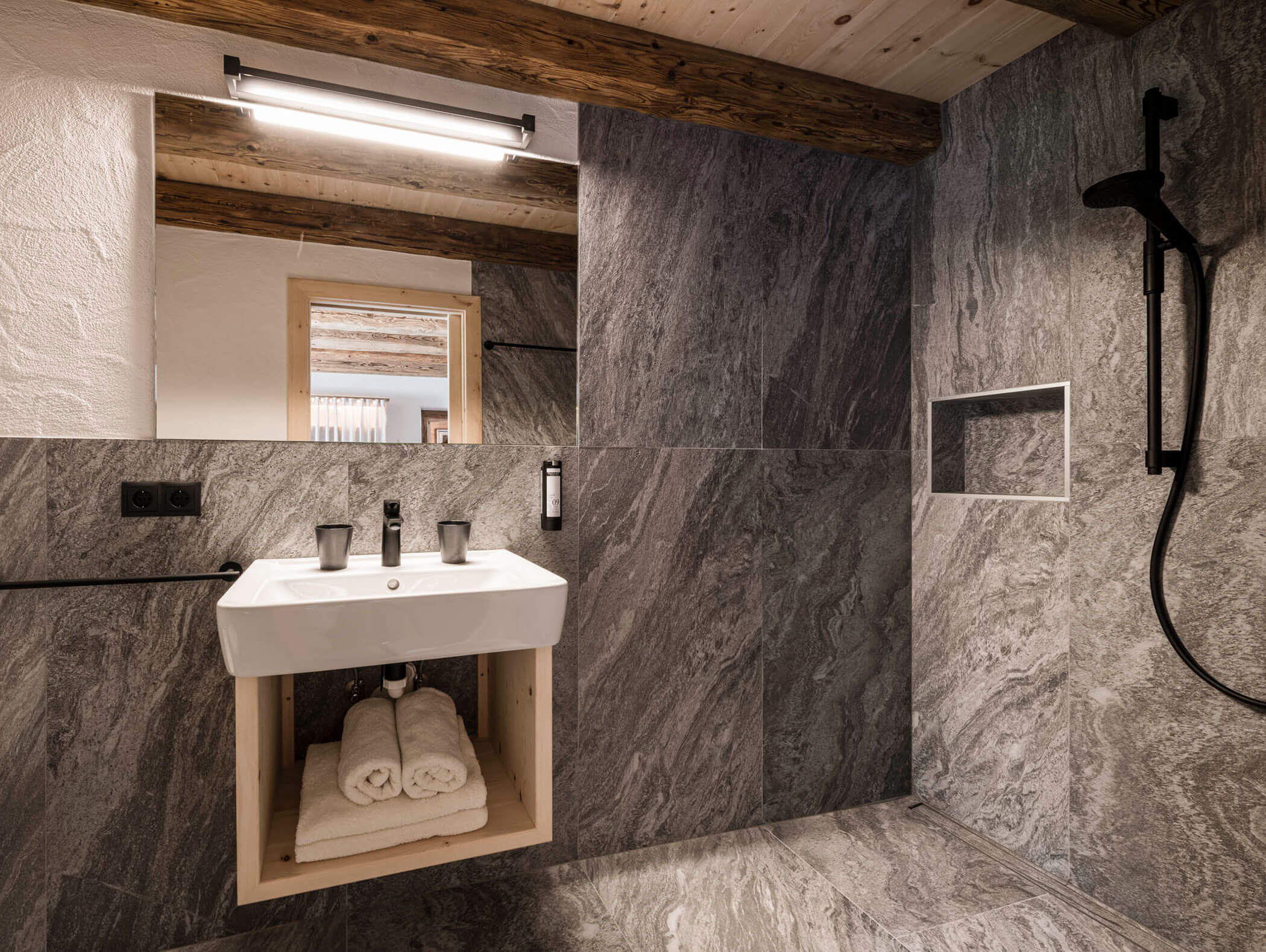 Modernes Badezimmer mit grauen Steinwänden, freiliegenden Holzbalken, weißem Waschbecken, Spiegel und gerollten Handtüchern darunter. - Maurlechnhof