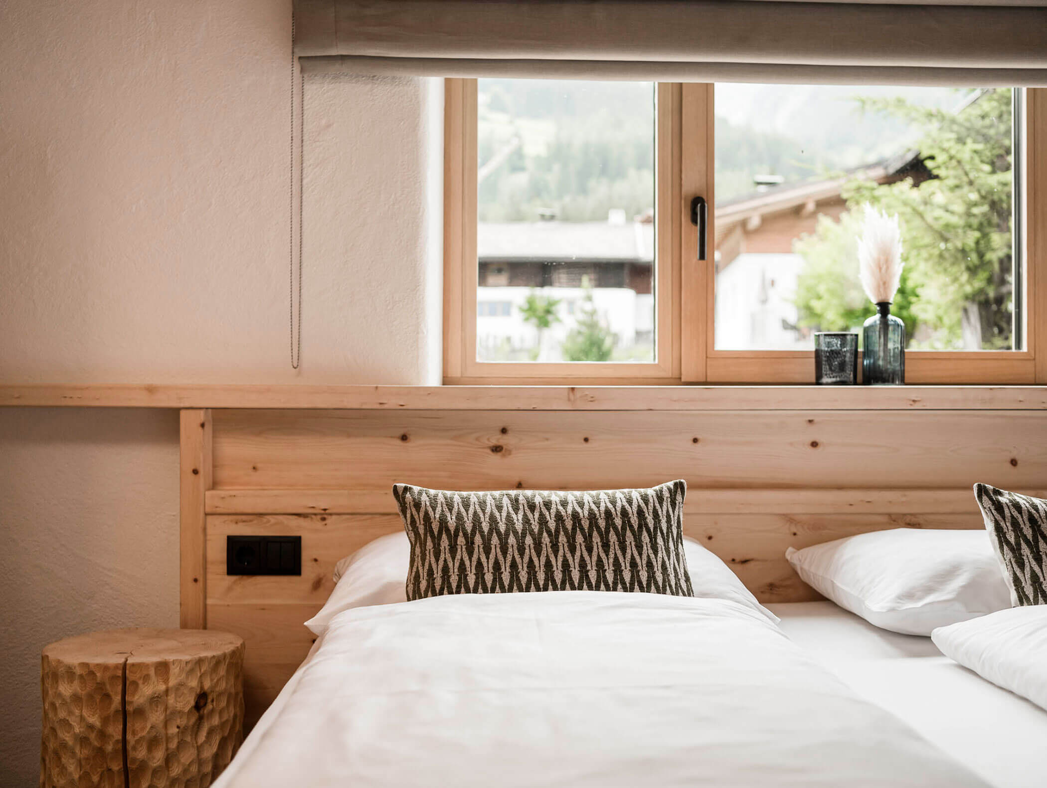 Ein gemütliches Bett mit weißer Bettwäsche neben einem Holzfenster, rustikales Dekor und Kissen in einem hellen Raum. - Maurlechnhof