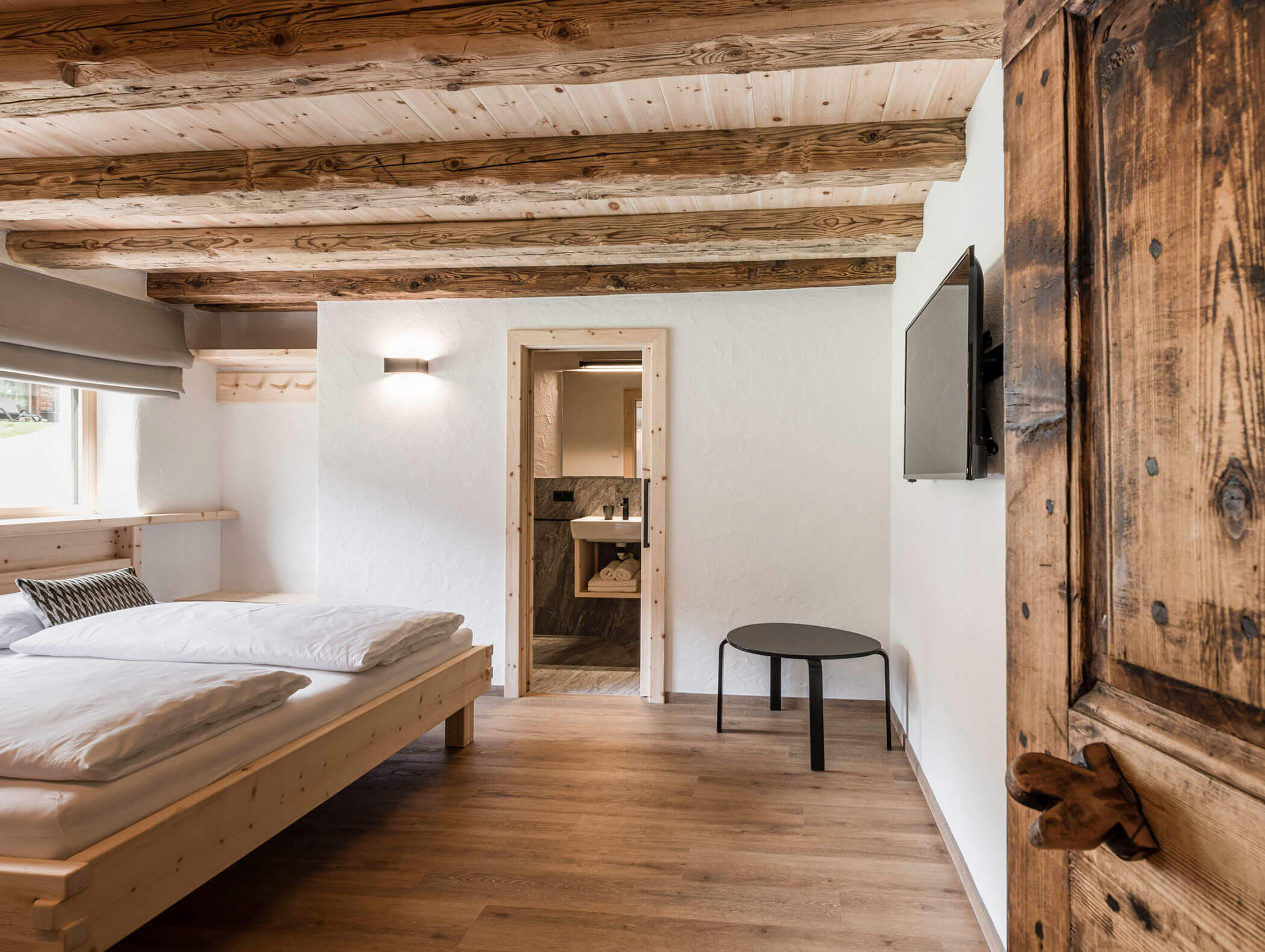 Gemütliches, rustikales Schlafzimmer mit Holzbalken, Doppelbett, TV und Blick in ein modern eingerichtetes Bad. - Maurlechnhof