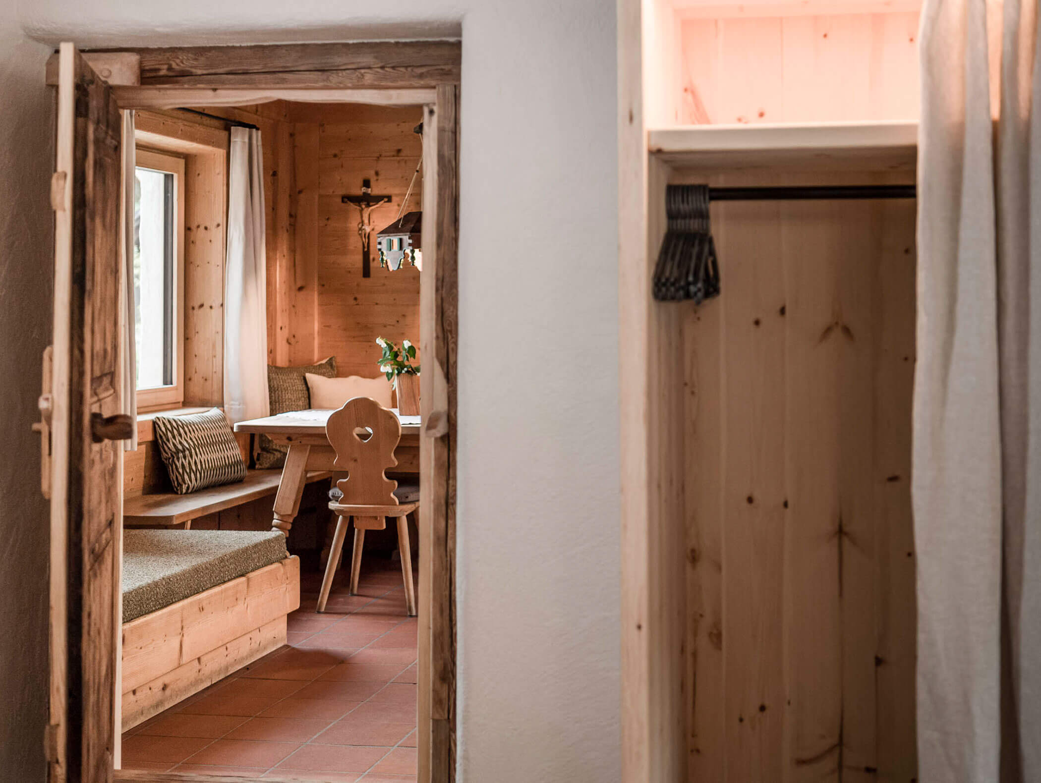 Gemütliche Essecke aus Holz mit einer Bank, einem Stuhl, einem Tisch, Kissen und einem Fenster, gesehen durch eine rustikale Türöffnung. - Maurlechnhof