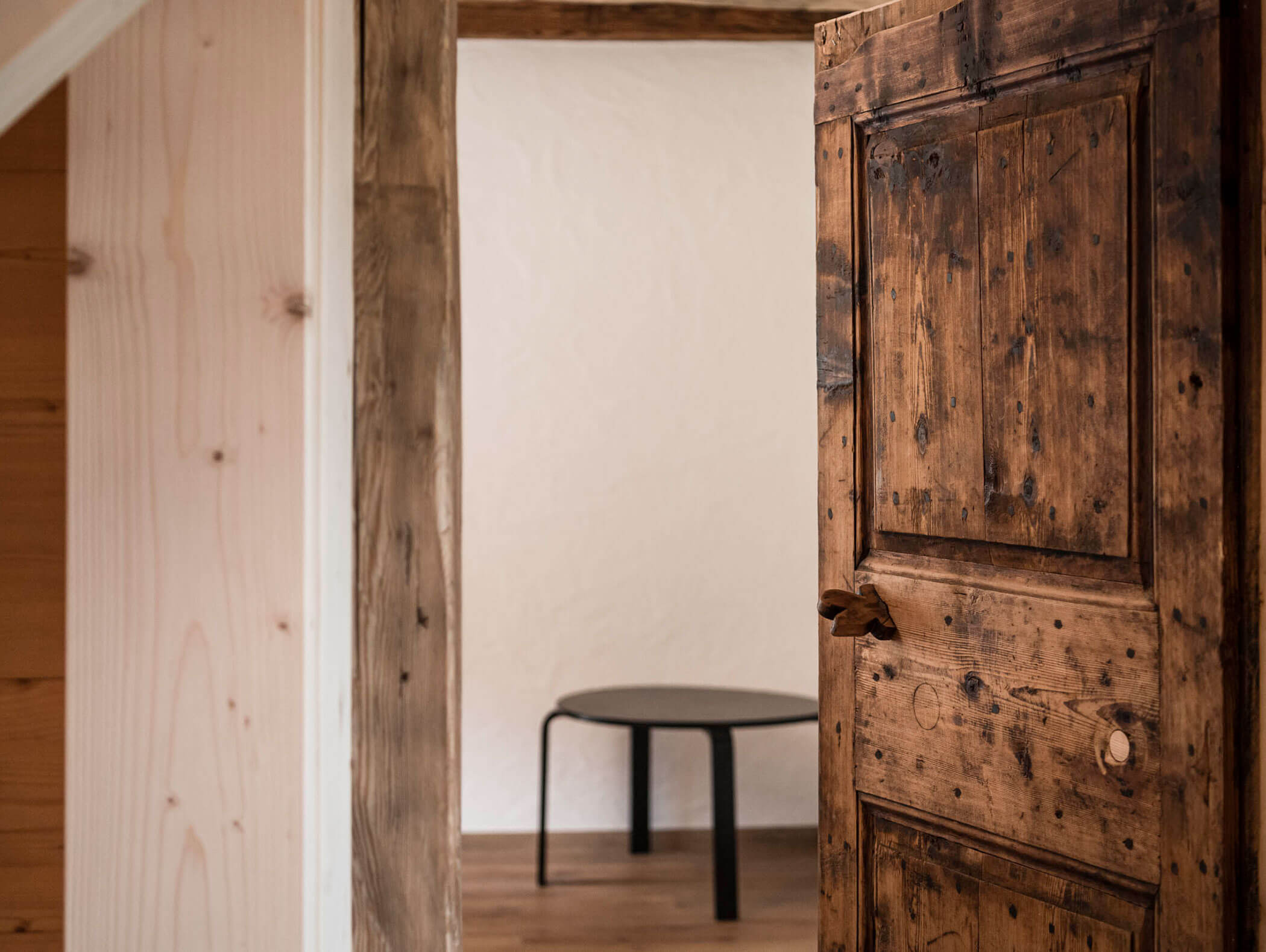 Eine rustikale Holztür führt in einen minimalistischen Raum mit einem runden schwarzen Tisch und Holzfußboden. - Maurlechnhof