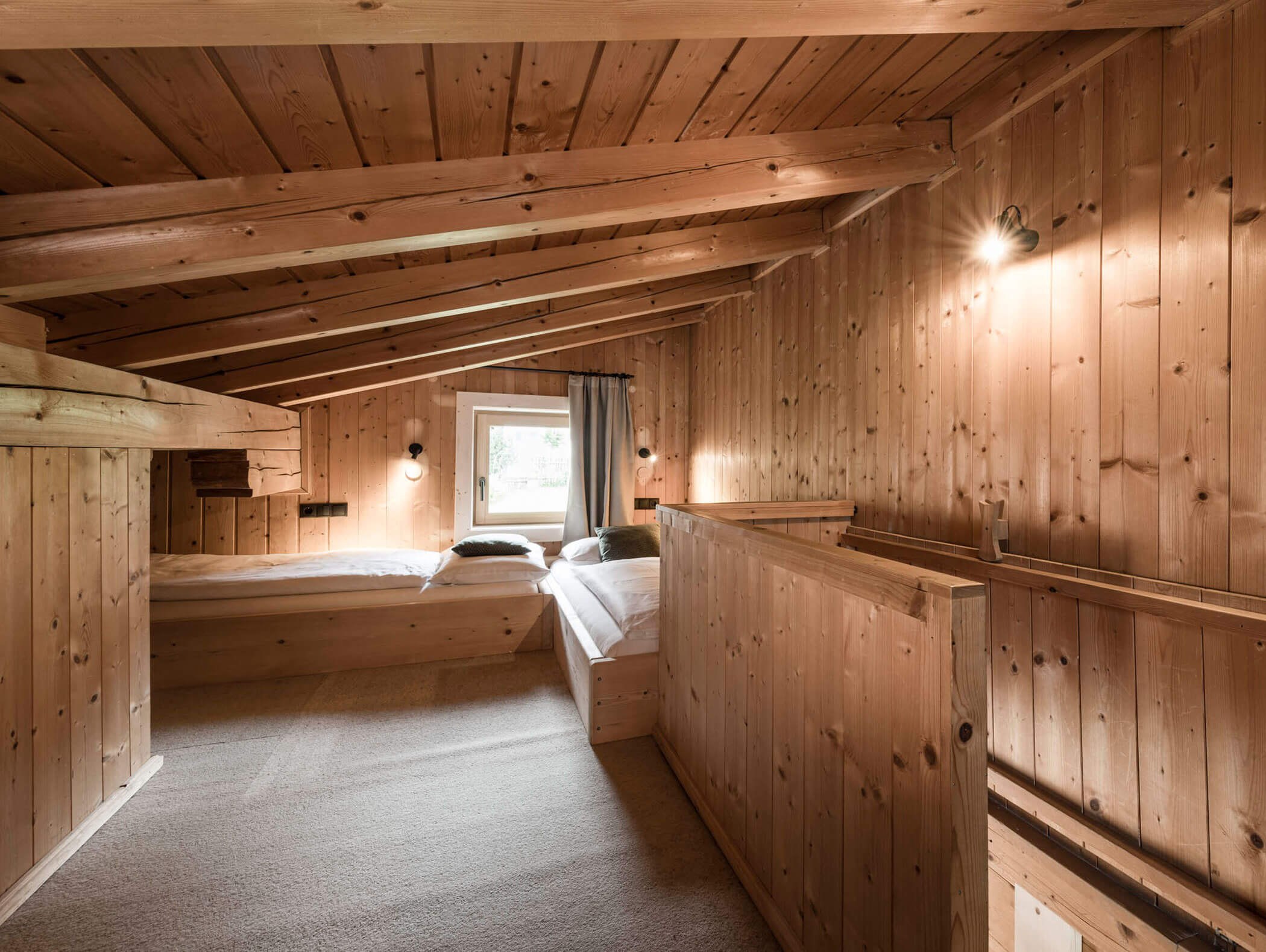 Gemütliches Loft-Zimmer aus Holz mit zwei Betten, schräger Decke, Wandlampen und einem Fenster mit grauen Vorhängen. - Maurlechnhof