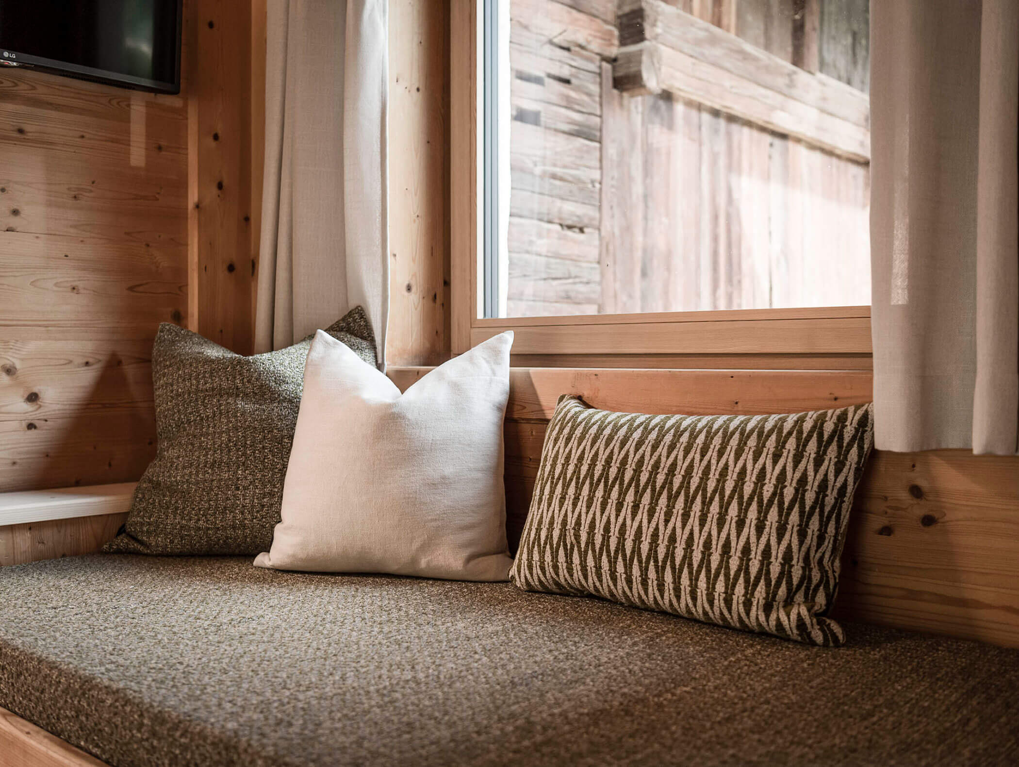 Ein gemütlicher Fensterplatz mit drei gemusterten Kissen in einem hölzernen Raum, Sonnenlicht scheint durch das Fenster. - Maurlechnhof