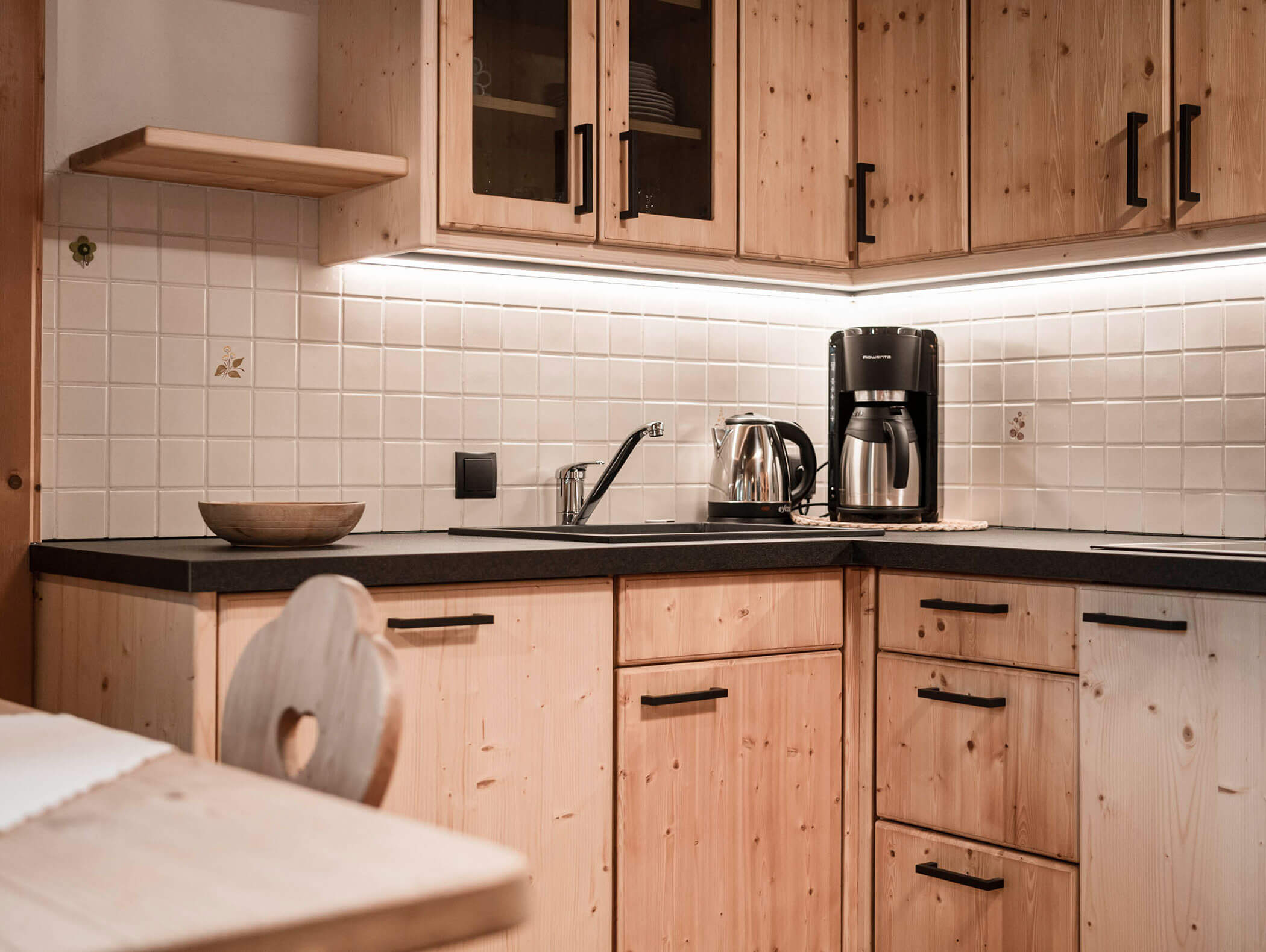 Gemütliche Holzküche mit schwarzer Arbeitsplatte, Kaffeemaschine, Wasserkocher, Spüle und gefliester Ablagefläche. - Maurlechnhof