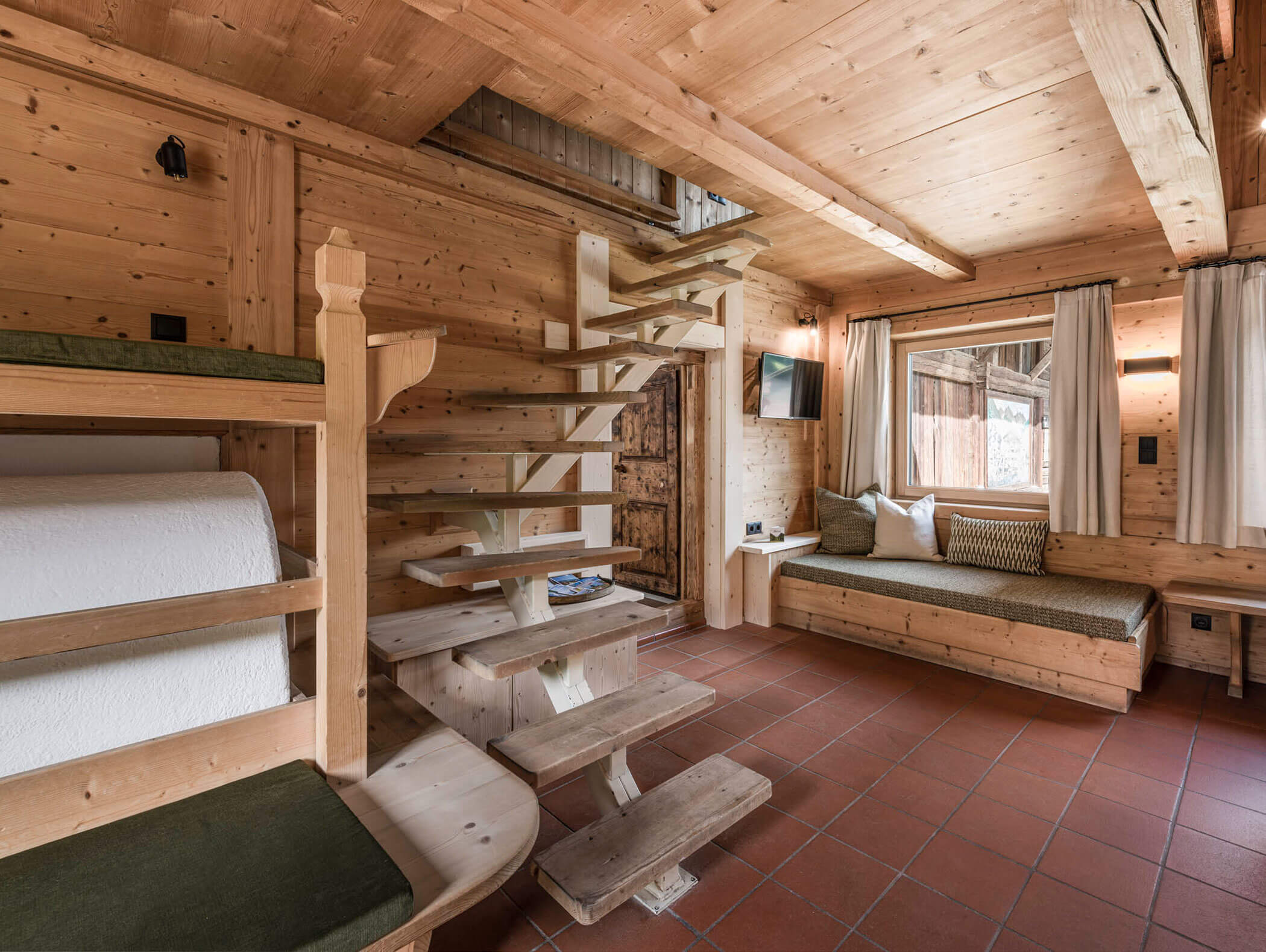 Gemütliche Holzeinrichtung mit offener Treppe, Etagenbett, Sofa, gefliestem Boden und Fenster mit Vorhängen. - Maurlechnhof