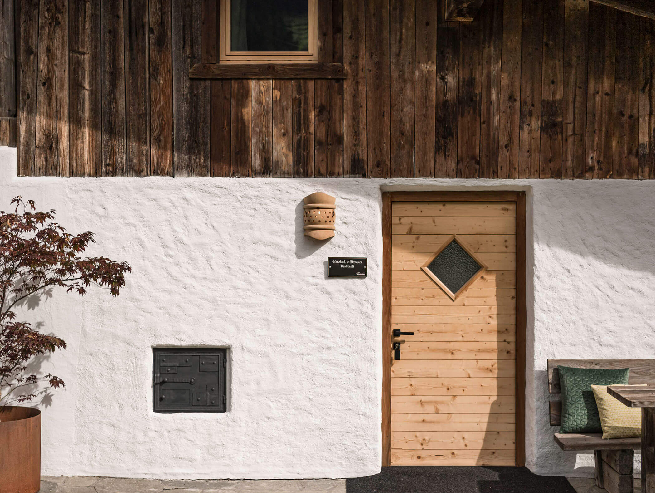 Holztür mit Rautenfenster in einer weißen Stuckwand, Bank und Topfbaum in der Nähe. - Maurlechnhof