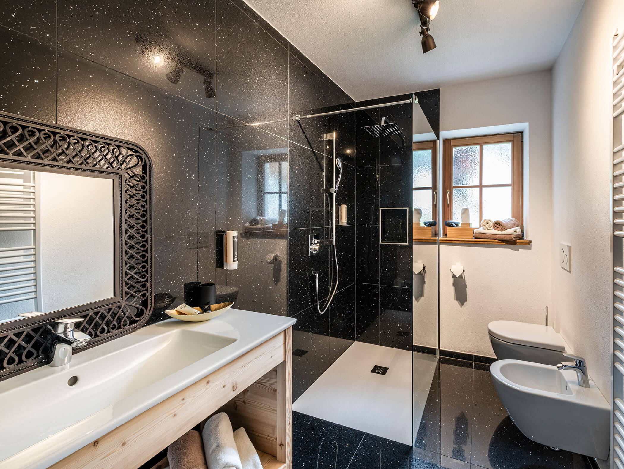 Modernes Badezimmer mit schwarz gefliester Dusche, weißem Waschbecken, Bidet, Toilette, großem Spiegel und Fenster mit Milchglas. - Maurlechnhof