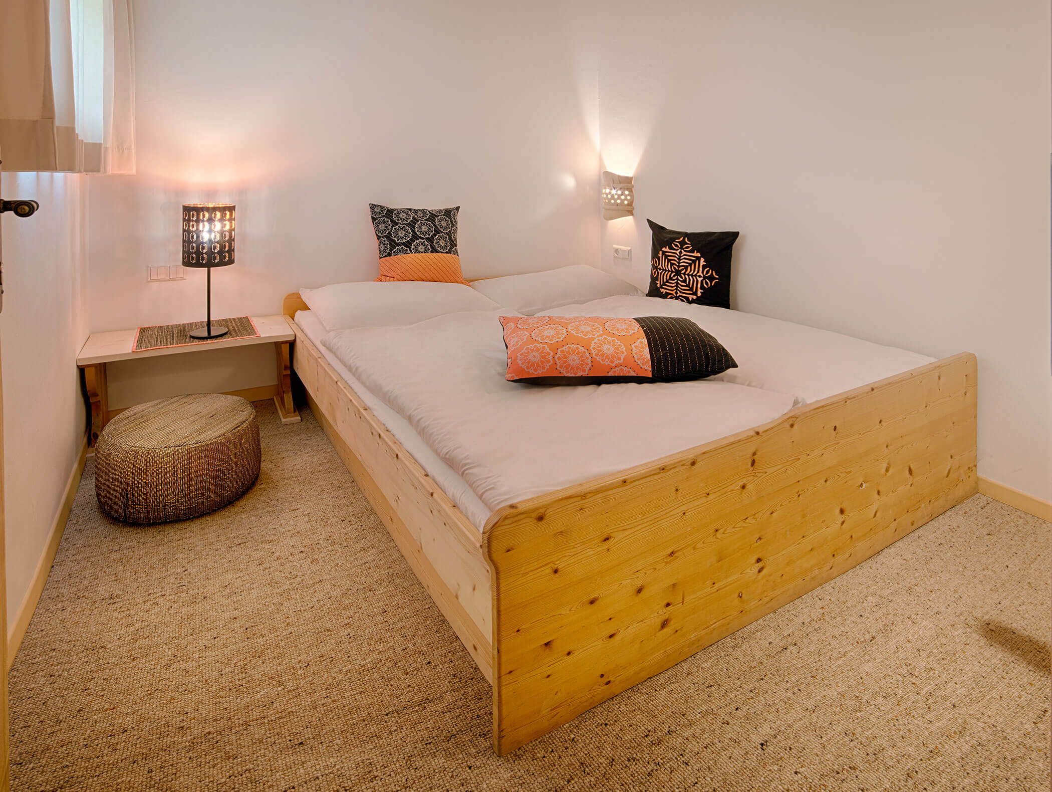 Gemütliches Schlafzimmer mit Holzbett, gemusterten Kissen, Nachttisch, Lampe und geflochtenem Hocker auf einem beigen Teppich. - Maurlechnhof