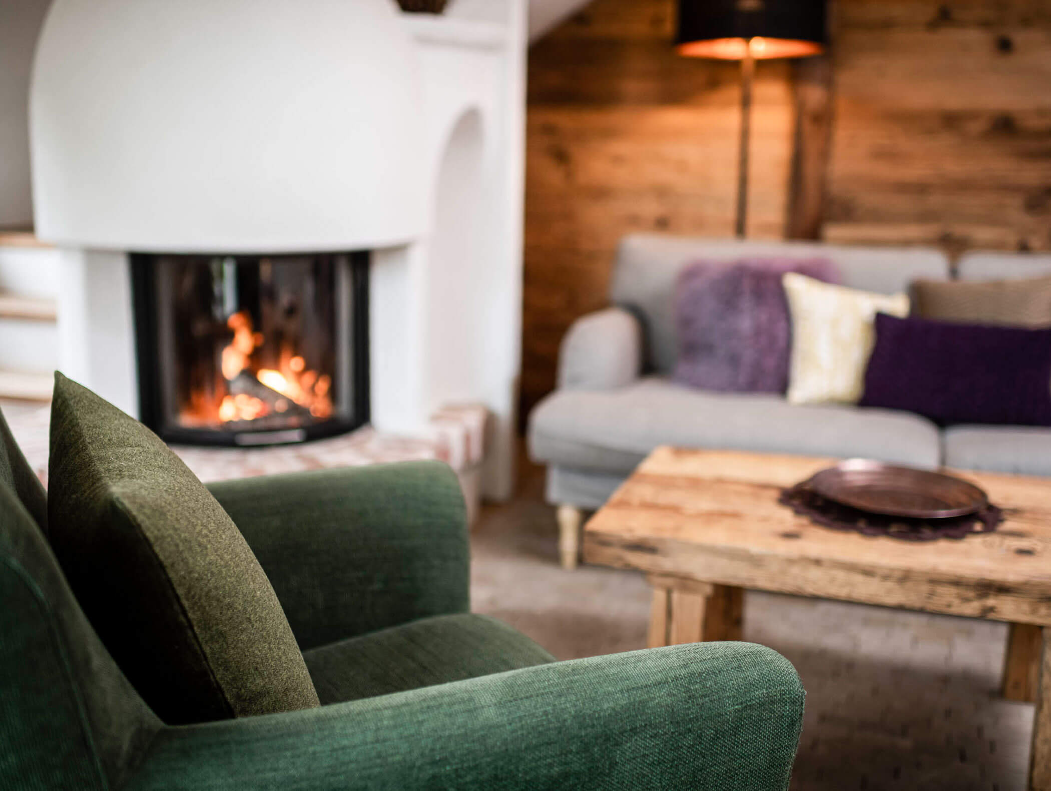 Gemütliches Wohnzimmer mit grünem Sessel, Holztisch, Couch und einem beleuchteten Kamin im Hintergrund. - Maurlechnhof