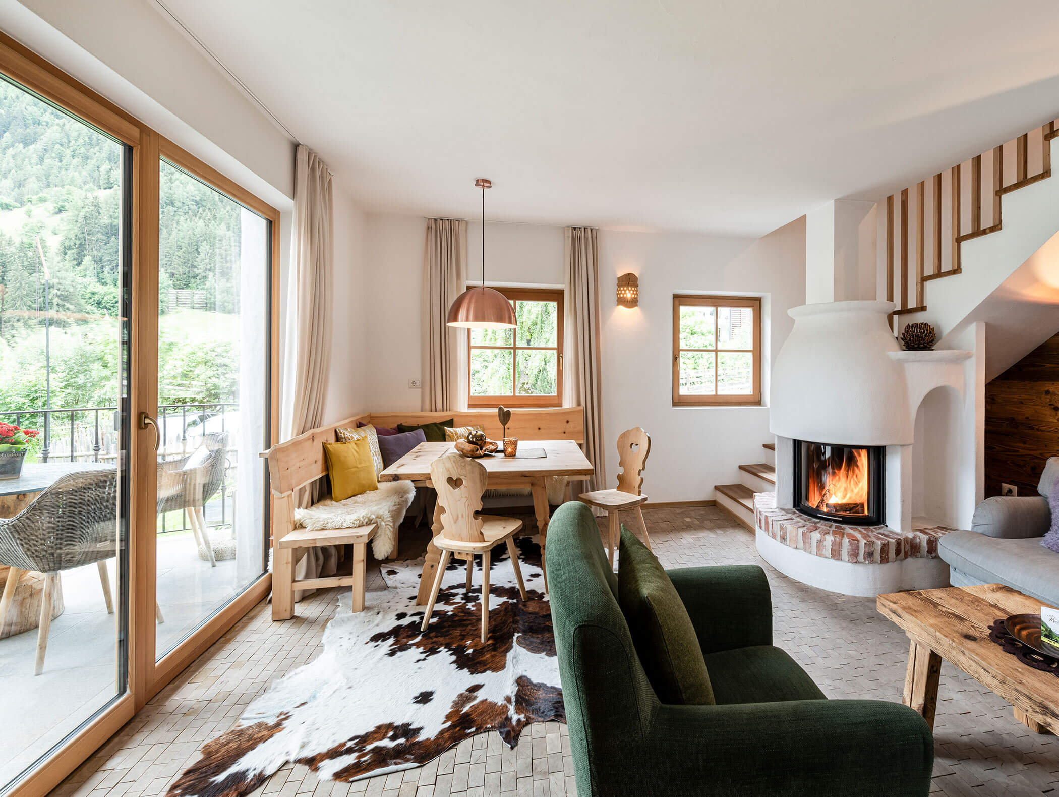 Gemütlicher Wohn- und Essbereich mit Kamin, Holzmöbeln, Fellteppich und großen Fenstern mit Blick ins Grüne. - Maurlechnhof