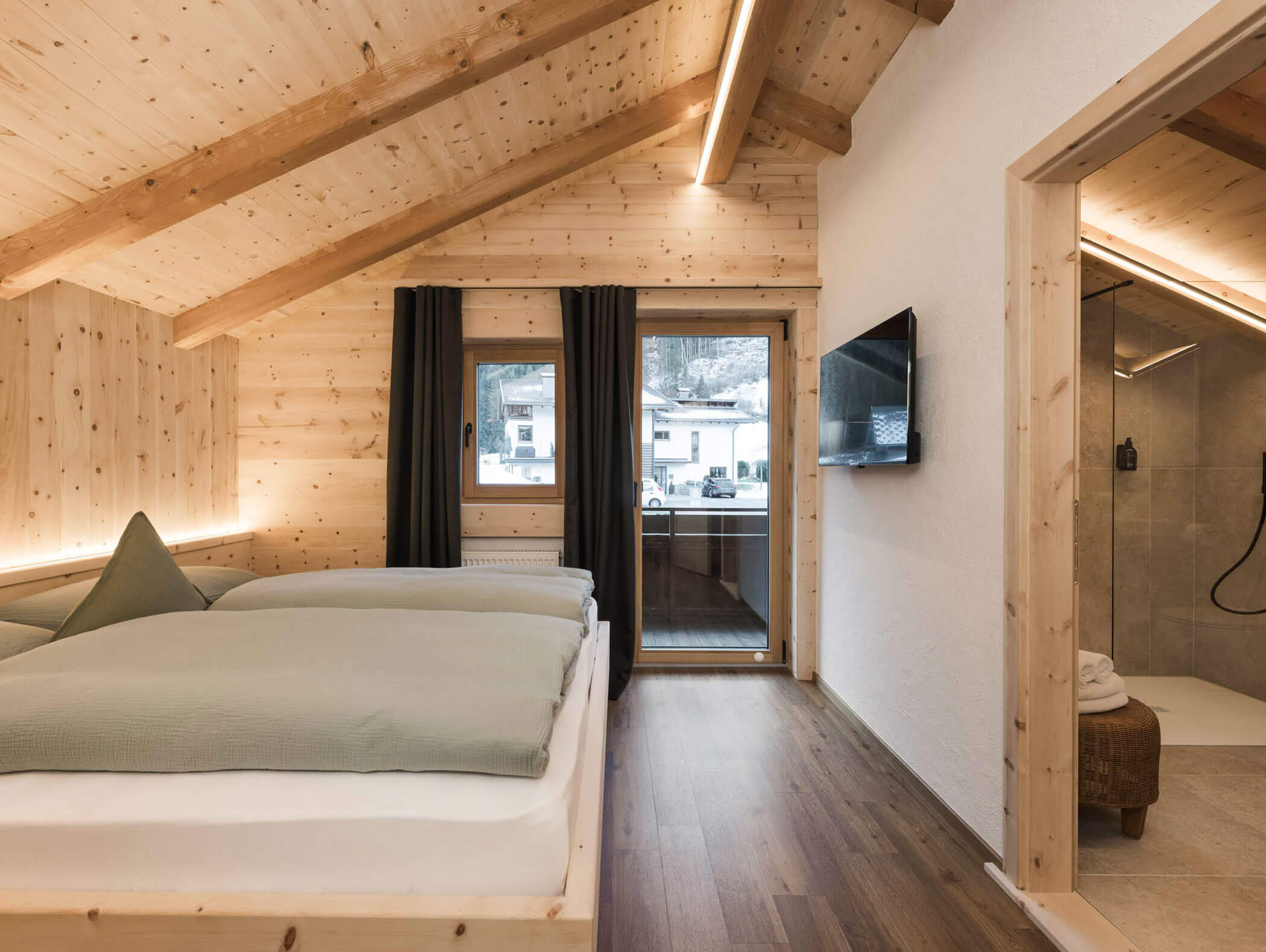 Gemütliches Holzschlafzimmer mit zwei Betten, einem an der Wand montierten Fernseher, einem großen Fenster und einem offenen Durchgang zu einer modernen Dusche. - Maurlechnhof