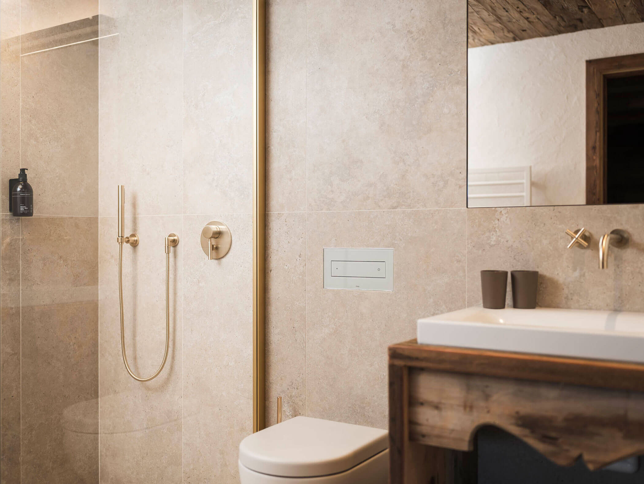 Modernes Badezimmer mit beigen Fliesen, goldenen Duscharmaturen, einer Toilette und einem hölzernen Waschtisch mit einem weißen Waschbecken. - Maurlechnhof