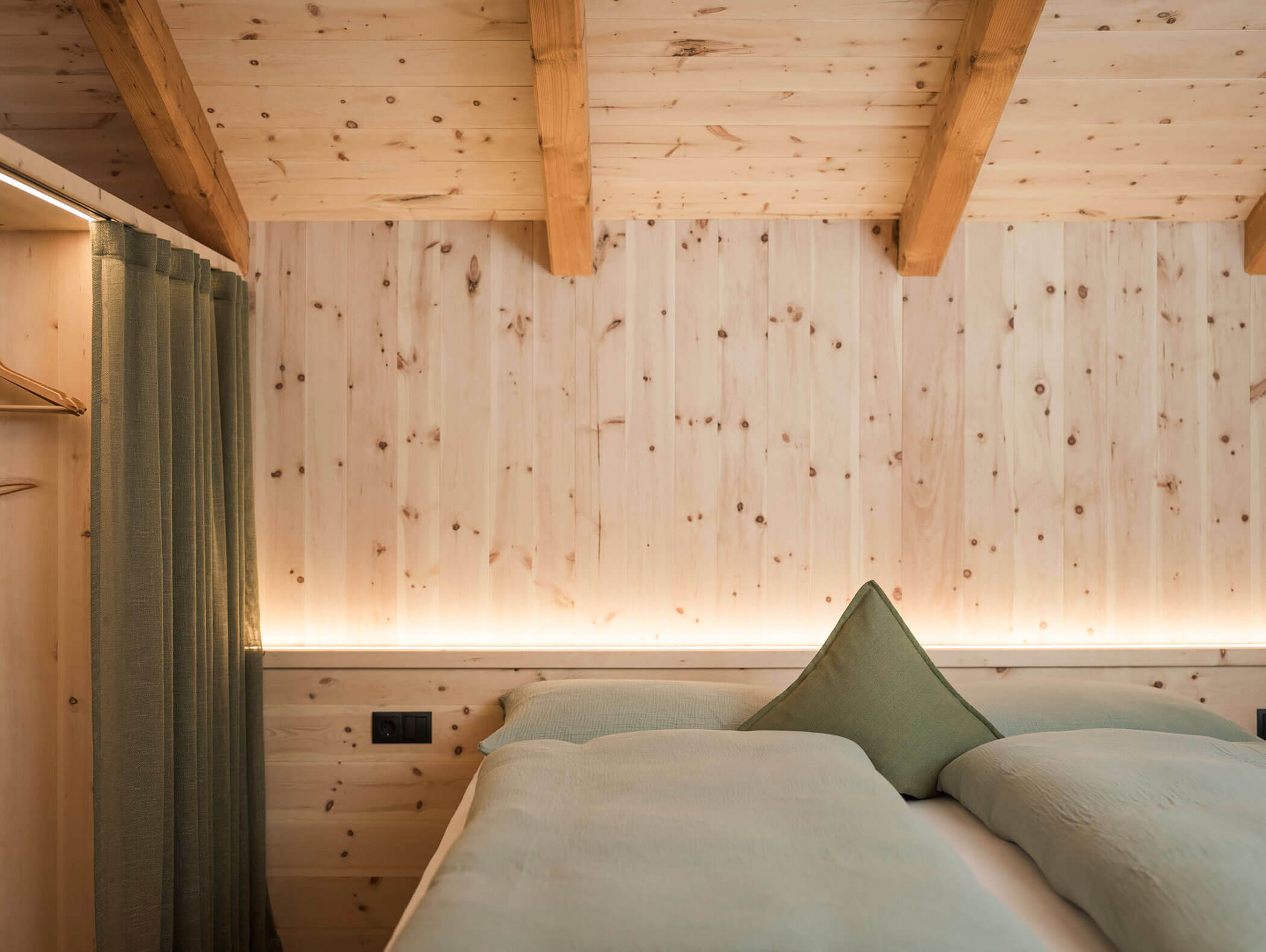 Minimalistisches Holzschlafzimmer mit freiliegenden Balken, sanfter Beleuchtung und ordentlich angeordneter grüner Bettwäsche und Vorhängen. - Maurlechnhof