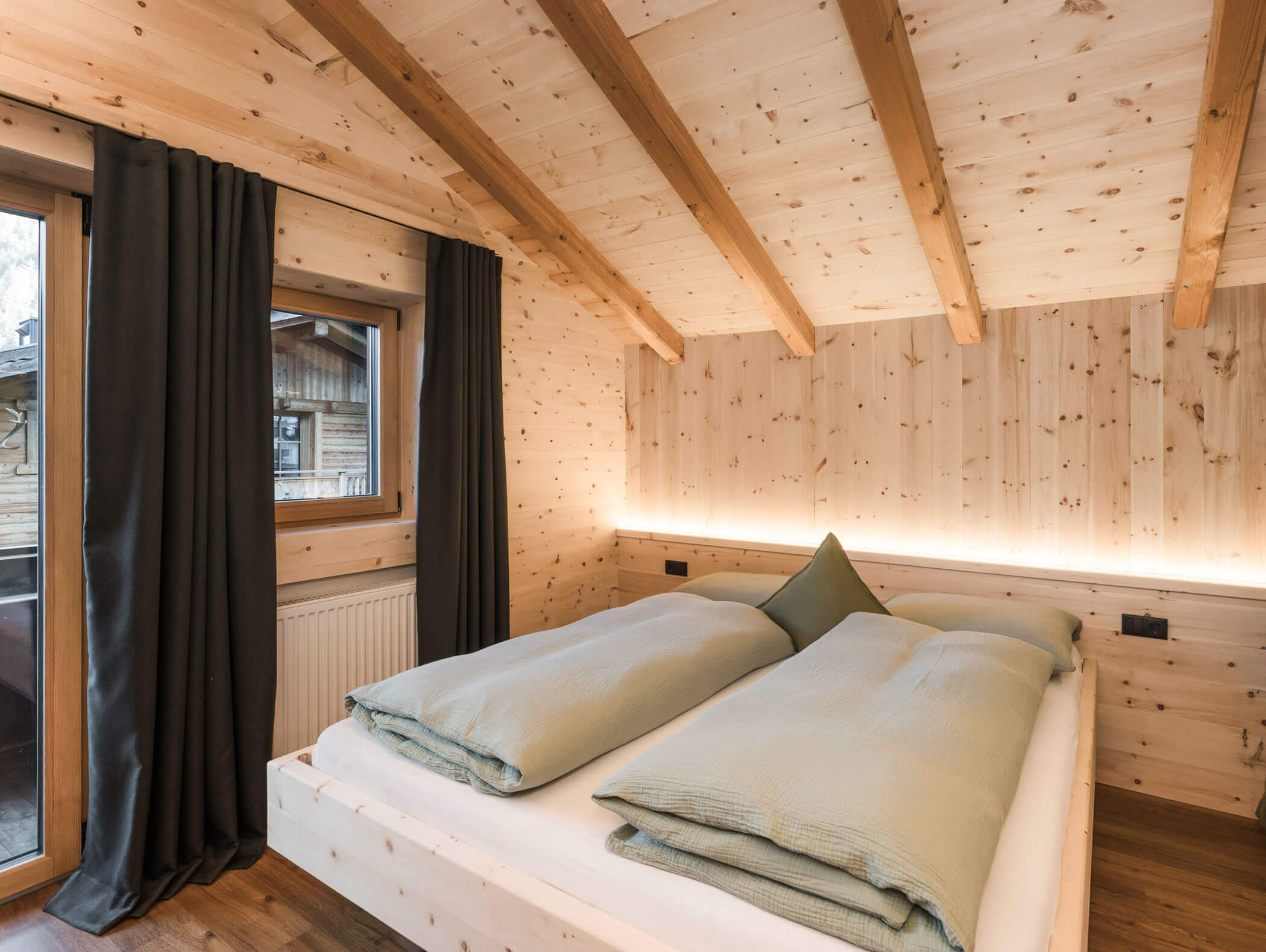 Gemütliches Schlafzimmer mit Holzwänden, Deckenbalken, großem Bett, grünem Bettzeug, Fenster und dunklen Vorhängen. - Maurlechnhof