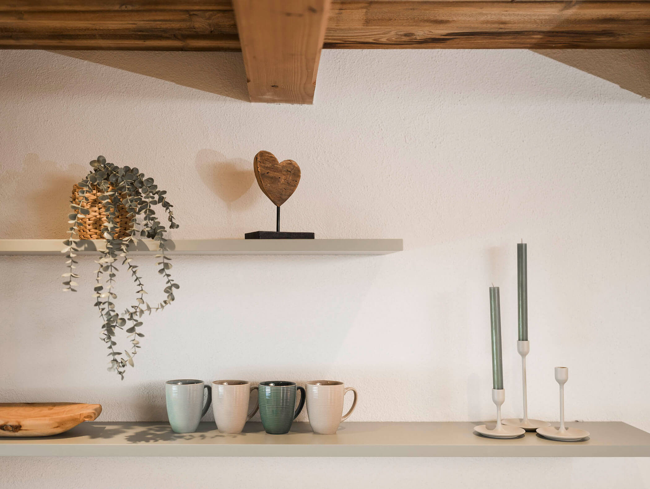 Zwei Regale mit Tassen, Kerzenständern, einem Holzherz, einer Pflanze und einer Holzschale an einer weißen Wand. - Maurlechnhof