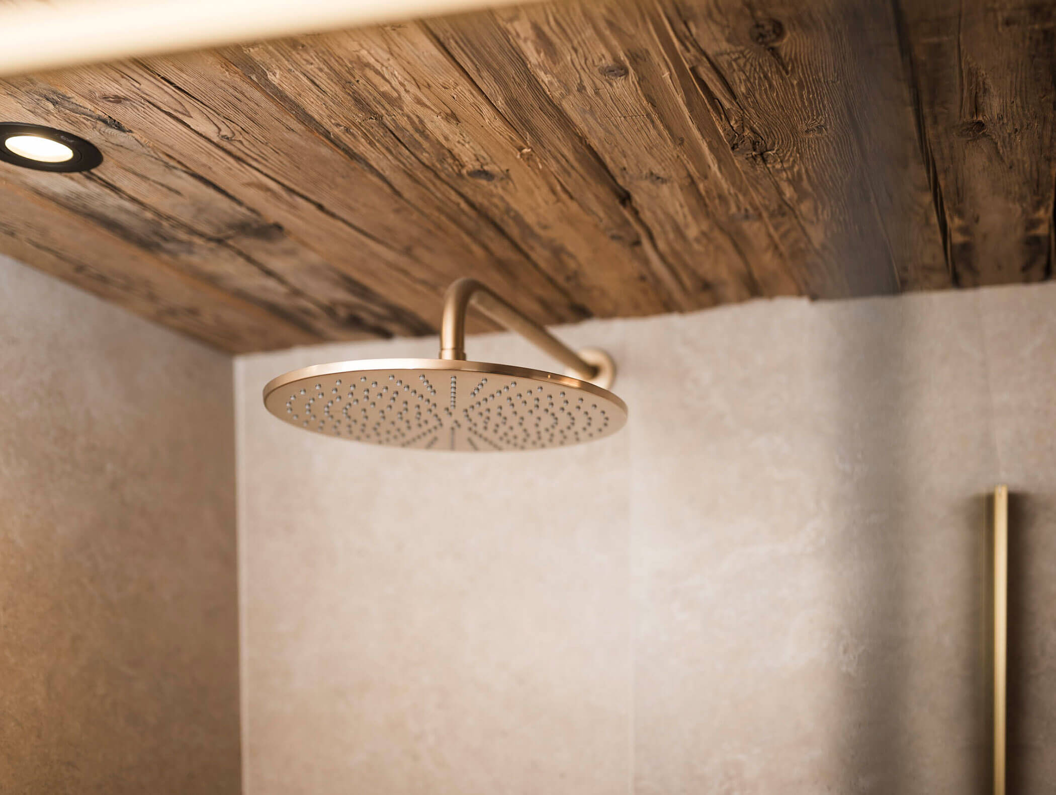 Goldfarbener Regen-Duschkopf an einer Holzdecke in einem beige gefliesten Badezimmer. - Maurlechnhof