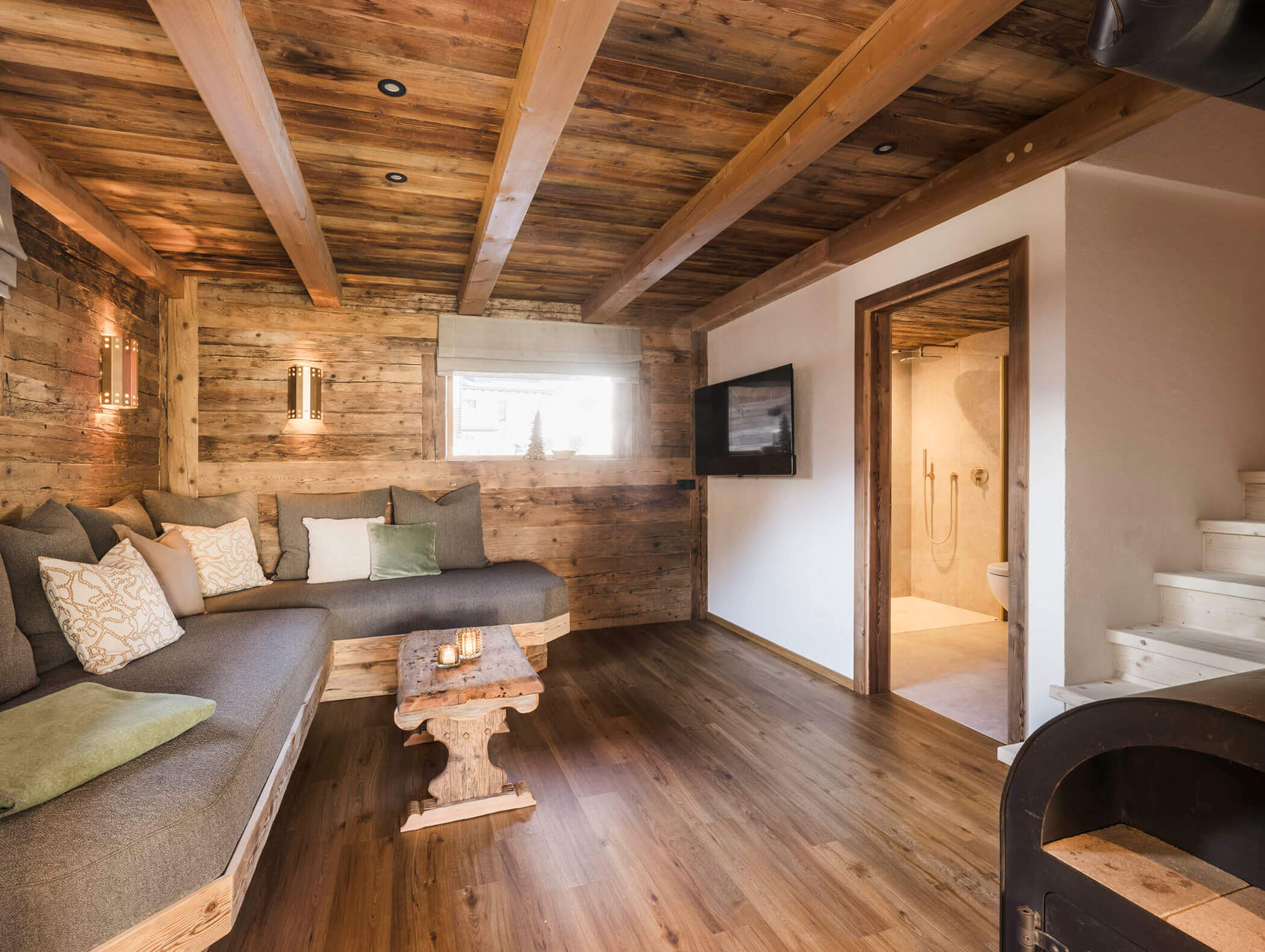Gemütliches, rustikales Wohnzimmer mit Holzwänden, -decke und -boden, einem grauen Sofa und einem Blick in ein modernes Badezimmer. - Maurlechnhof