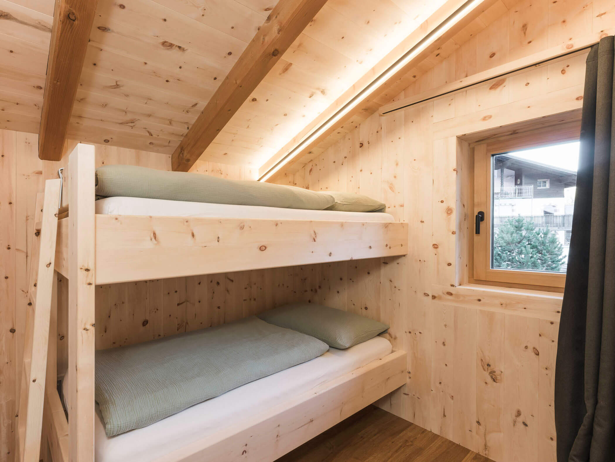 Gemütliches Holzzimmer mit Etagenbett am Fenster, unter einer schrägen Decke mit sichtbaren Balken. - Maurlechnhof
