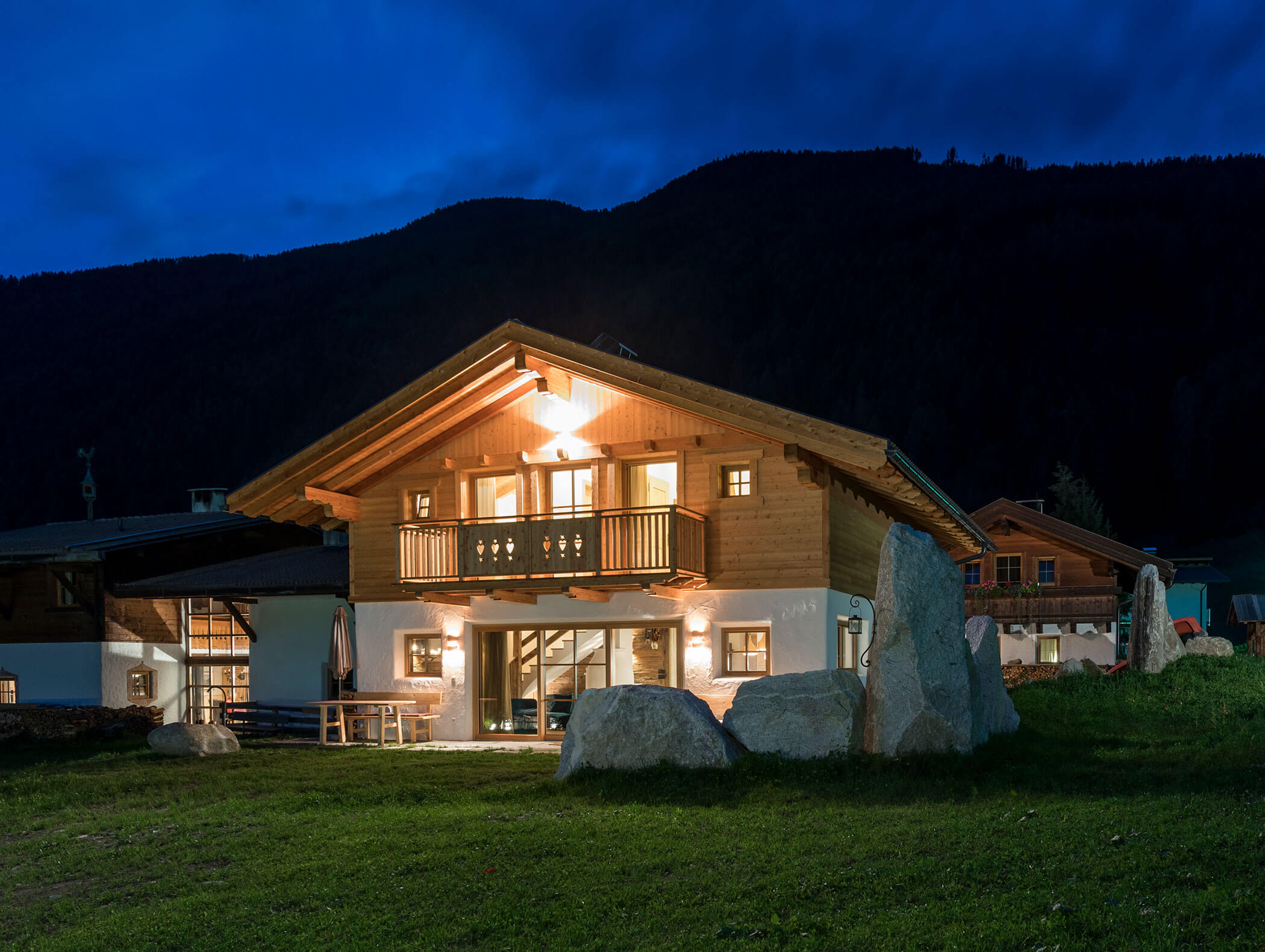 Ein gemütliches, beleuchtetes Holzchalet steht nachts auf einem grasbewachsenen Hof mit den Silhouetten der Berge im Hintergrund. - Maurlechnhof