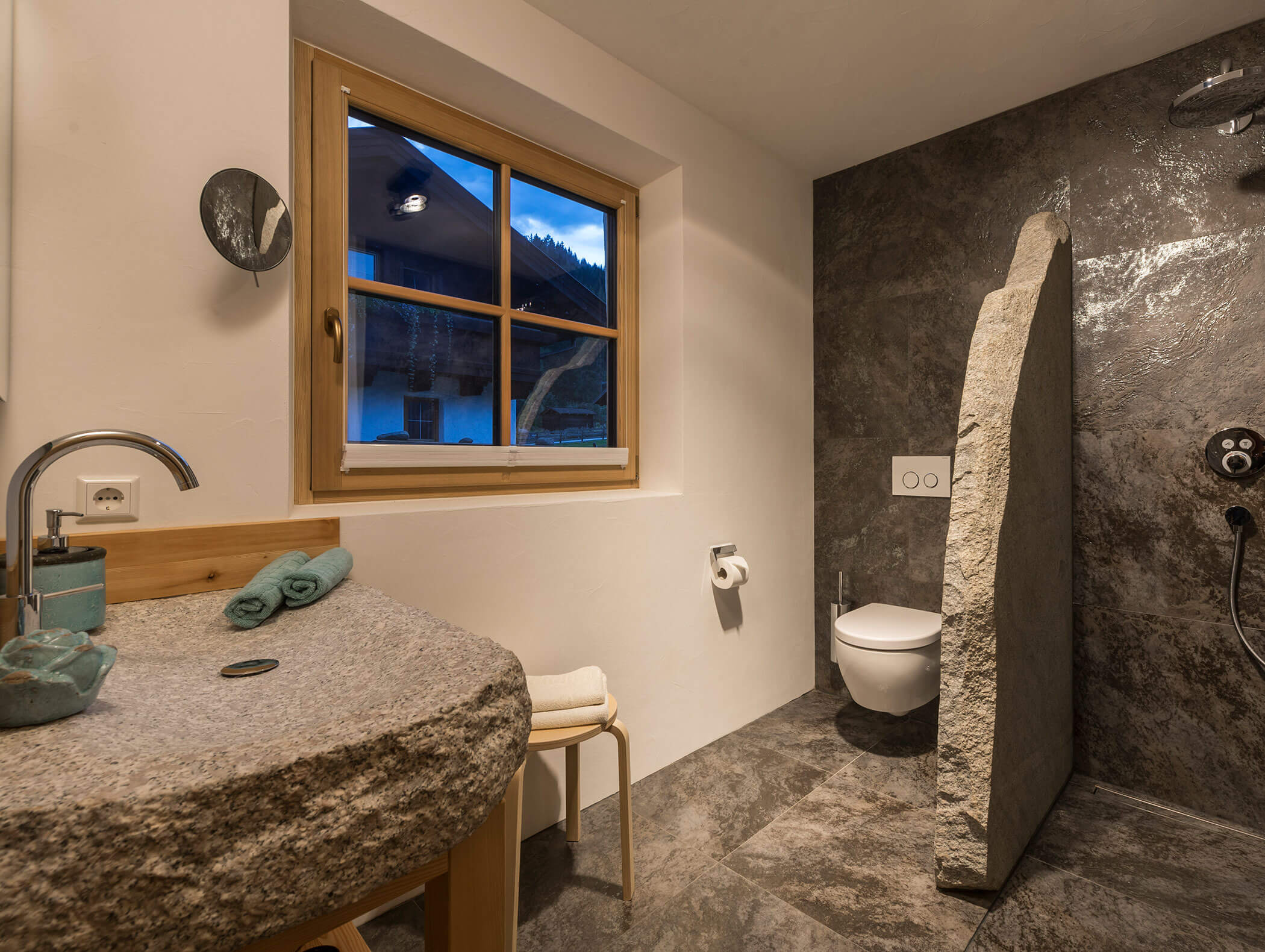 Modernes Badezimmer mit Steinwaschbecken und -dusche, wandmontierter Toilette, Holzfenster und grau gefliestem Boden und Wänden. - Maurlechnhof