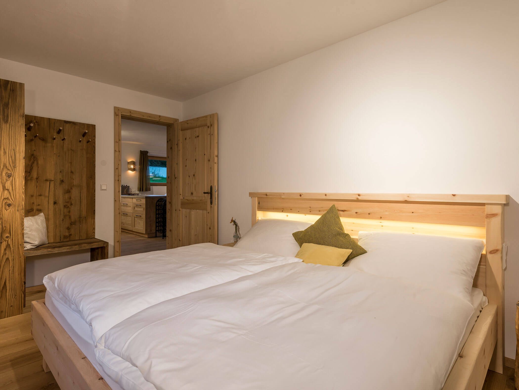 Gemütliches Schlafzimmer mit Holzbett, weißem Bettzeug und zwei Kissen zur Akzentuierung; Tür zum Küchenbereich offen. - Maurlechnhof