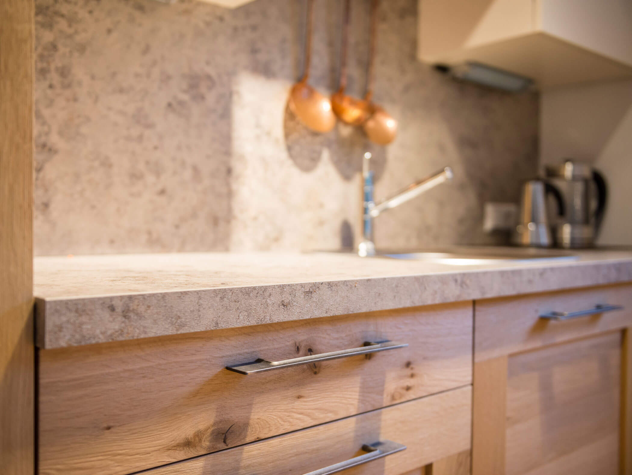 Nahaufnahme einer modernen Küchenarbeitsplatte mit Holzschränken, Wasserkocher, Spülbecken und hängenden Utensilien im Hintergrund. - Maurlechnhof