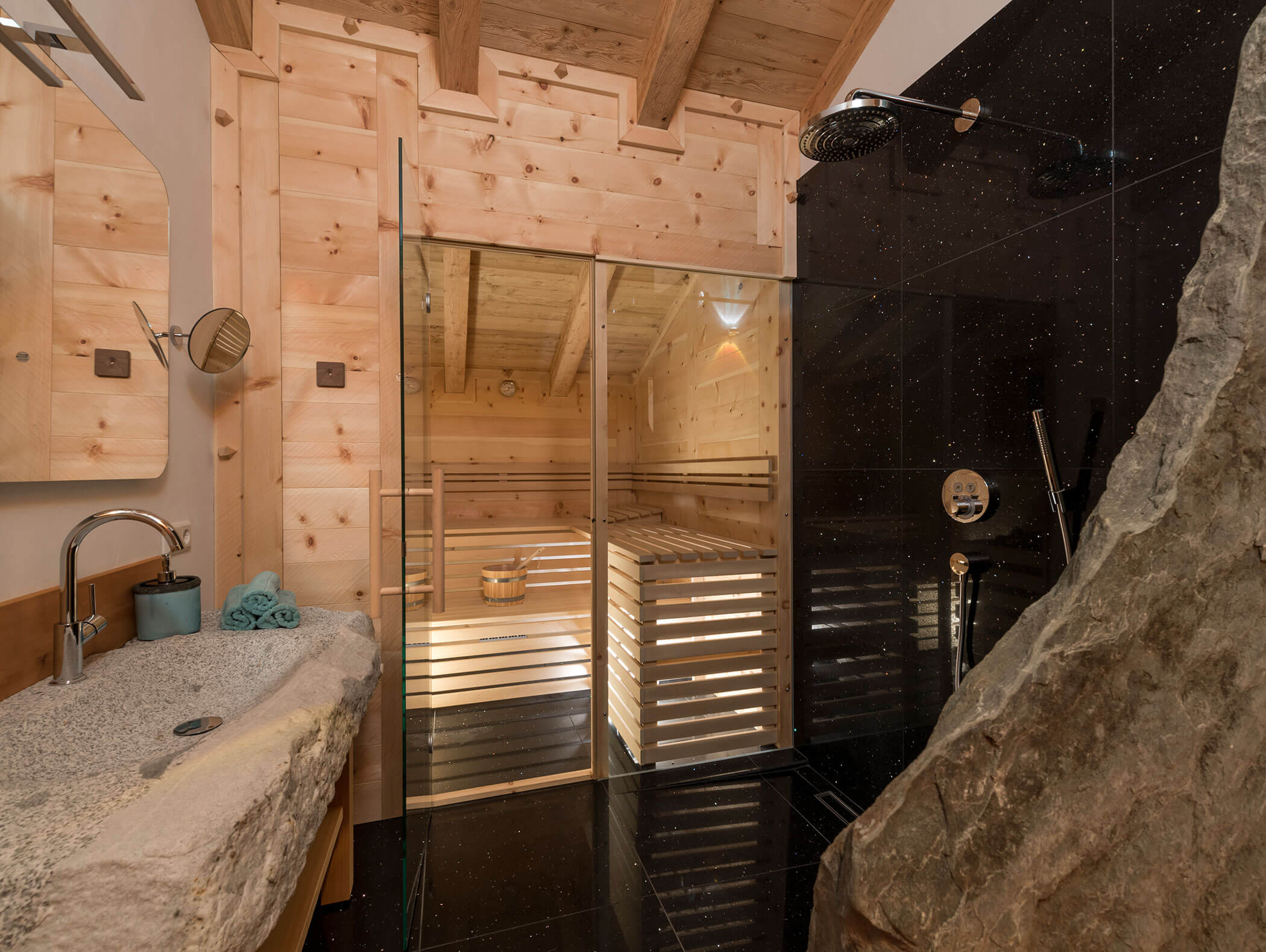 Modernes Badezimmer mit Steinwaschbecken, Glasdusche, dunklen Fliesen und einer durch eine Glastür sichtbaren Holzsauna. - Maurlechnhof