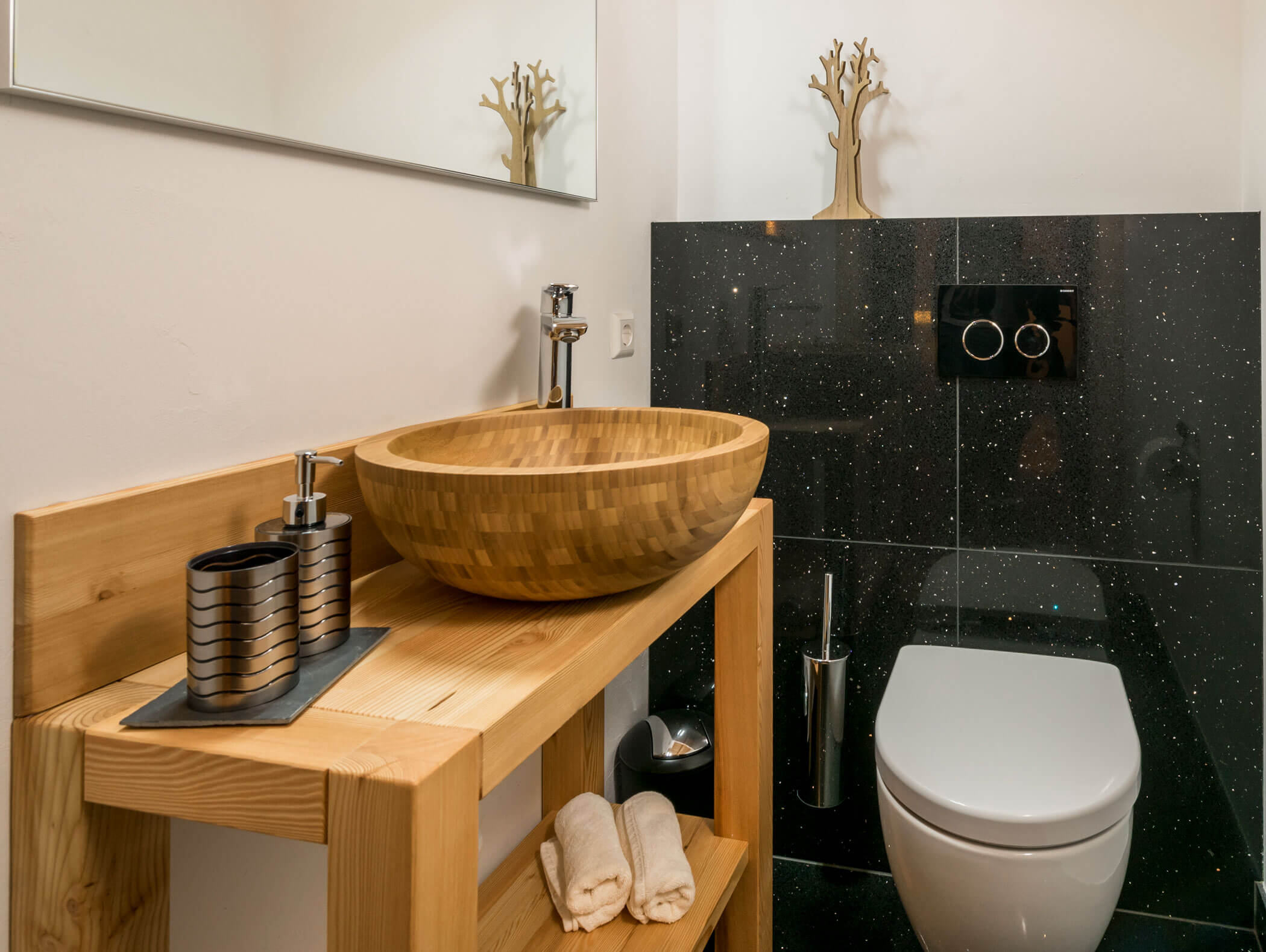 Modernes Badezimmer mit einem Waschbecken aus Holz, einer schwarzen Arbeitsplatte, gerollten Handtüchern und dekorativem Baumschmuck. - Maurlechnhof