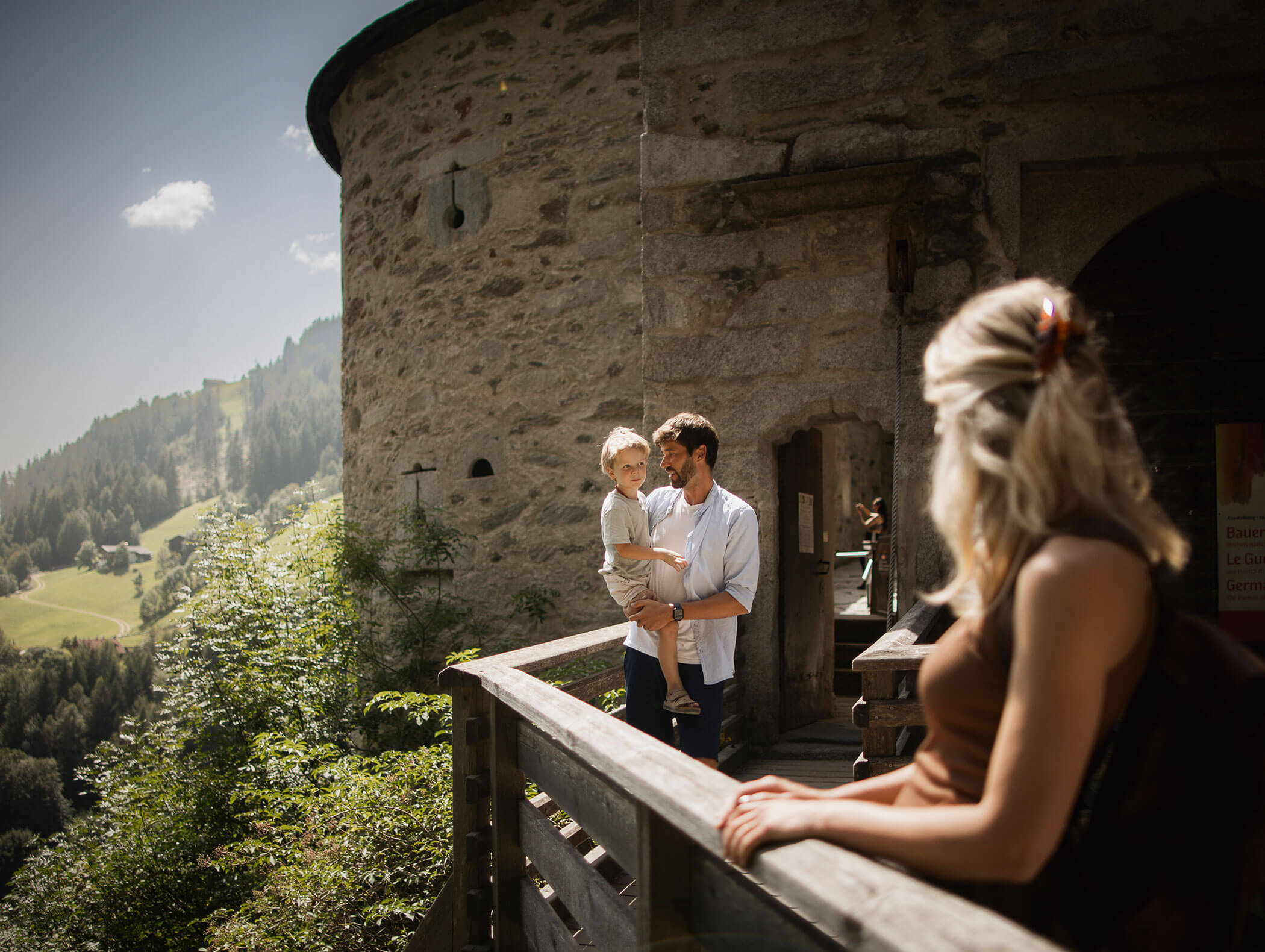 Ein Mann hält ein Kind auf einem steinernen Balkon, während eine Frau zuschaut; Berge und Bäume im Hintergrund. - Maurlechnhof