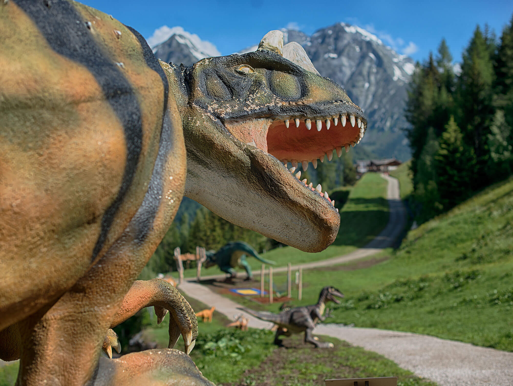 Eine große Dinosaurier-Skulptur mit offenen Kiefern steht auf einem grasbewachsenen Weg mit Bergen im Hintergrund. - Maurlechnhof