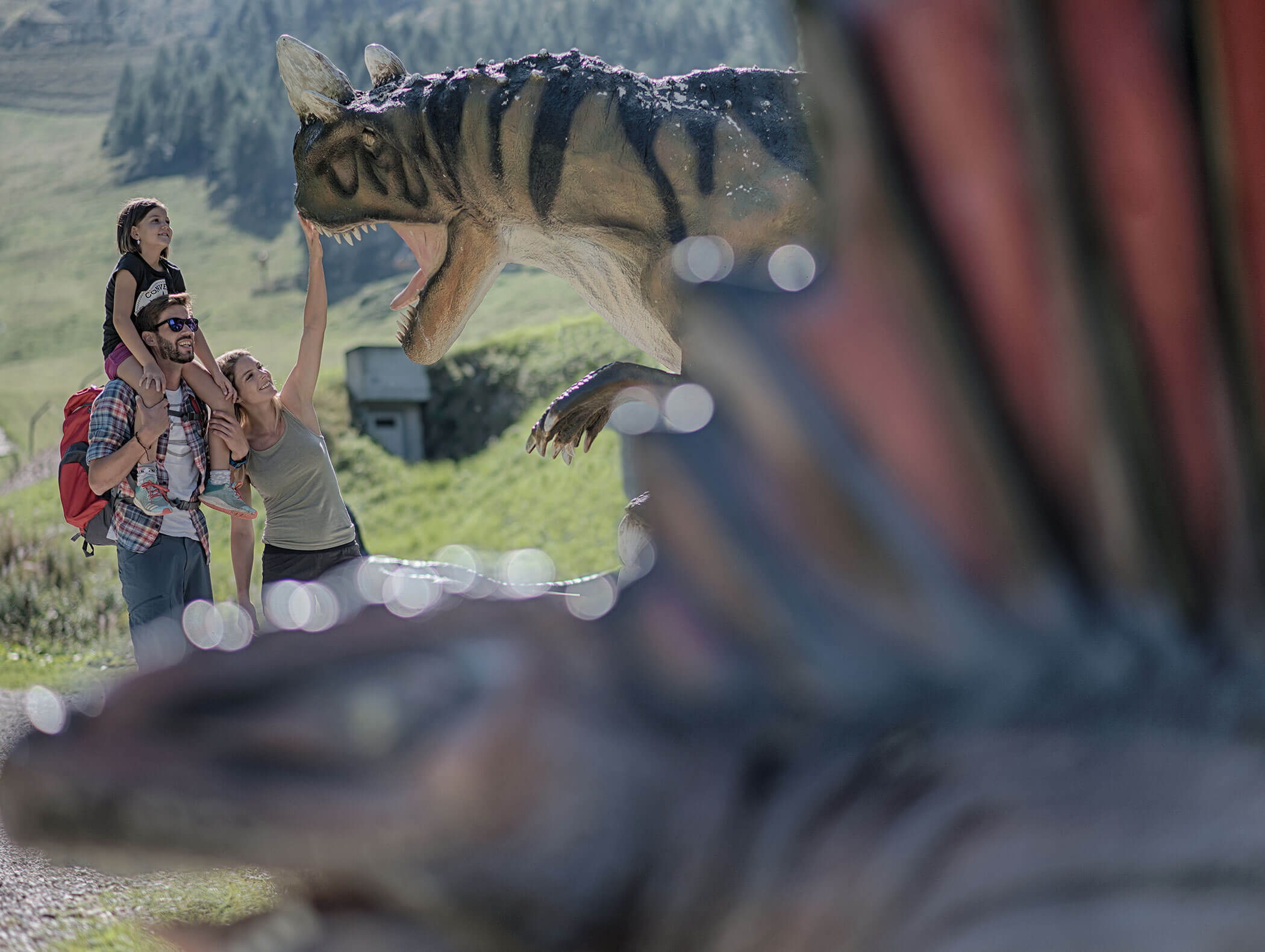 Eine Familie interagiert an einem sonnigen Tag im Freien mit großen Dinosaurierstatuen, umgeben von Grün und Bergen. - Maurlechnhof