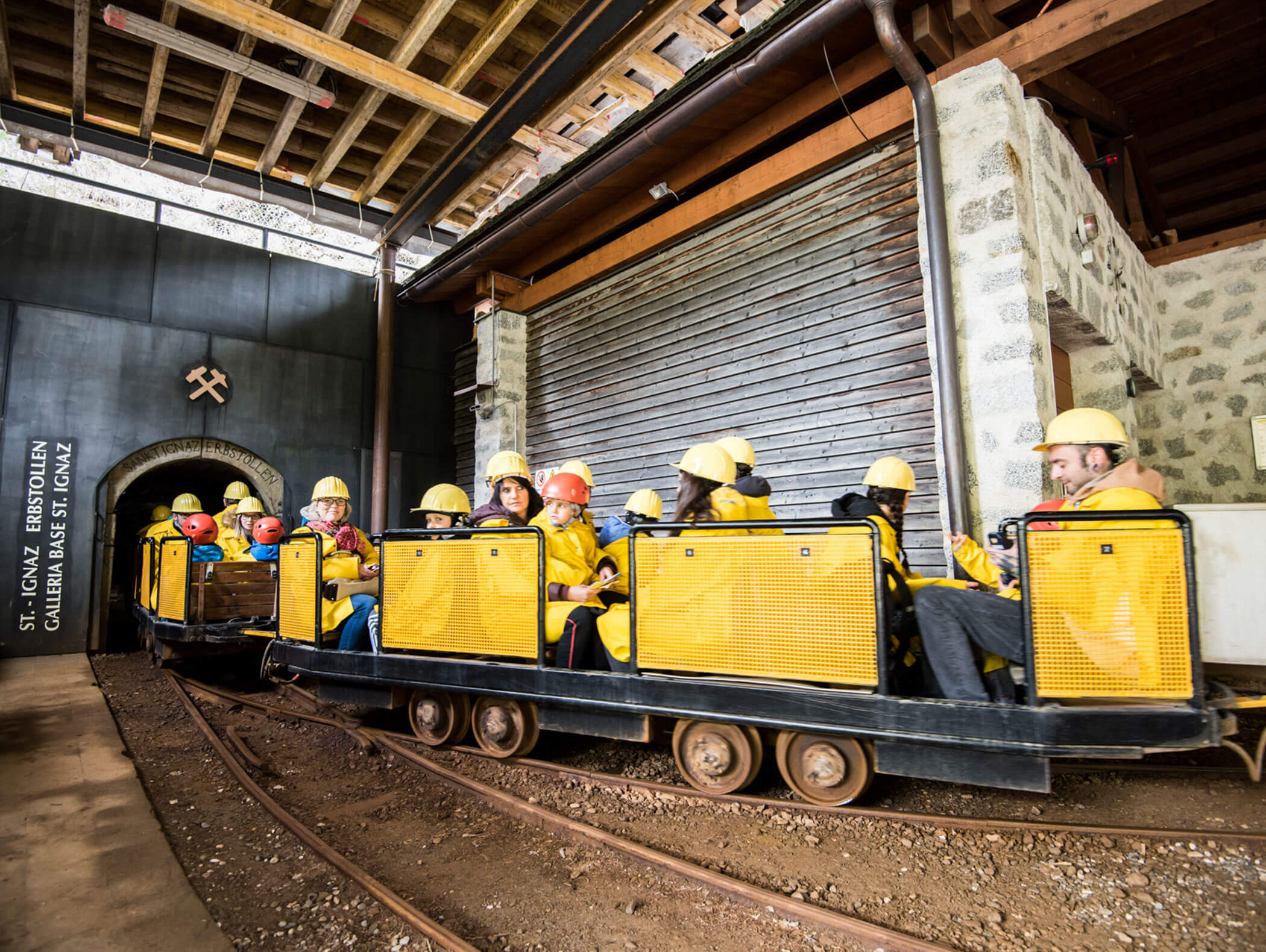Menschen mit Schutzhelmen sitzen in einem gelben Grubenzug, der in einen Tunnel innerhalb eines Industriegebäudes einfährt. - Maurlechnhof