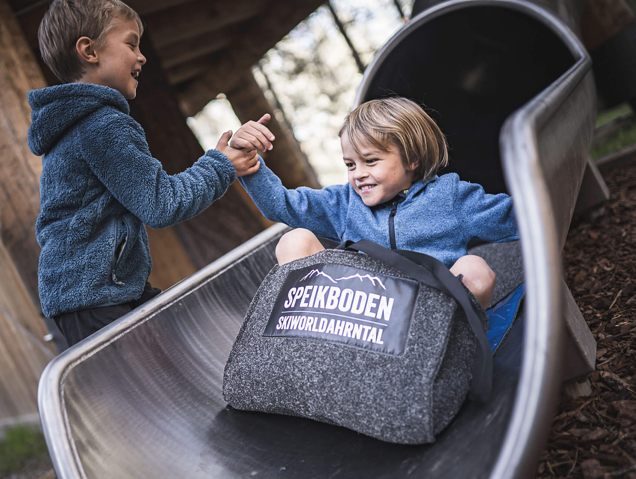 Zwei glückliche Kinder spielen auf einer Rutsche, wobei das eine dem anderen mit einem Speikboden-Skiworld-Sack hochhilft. - Maurlechnhof