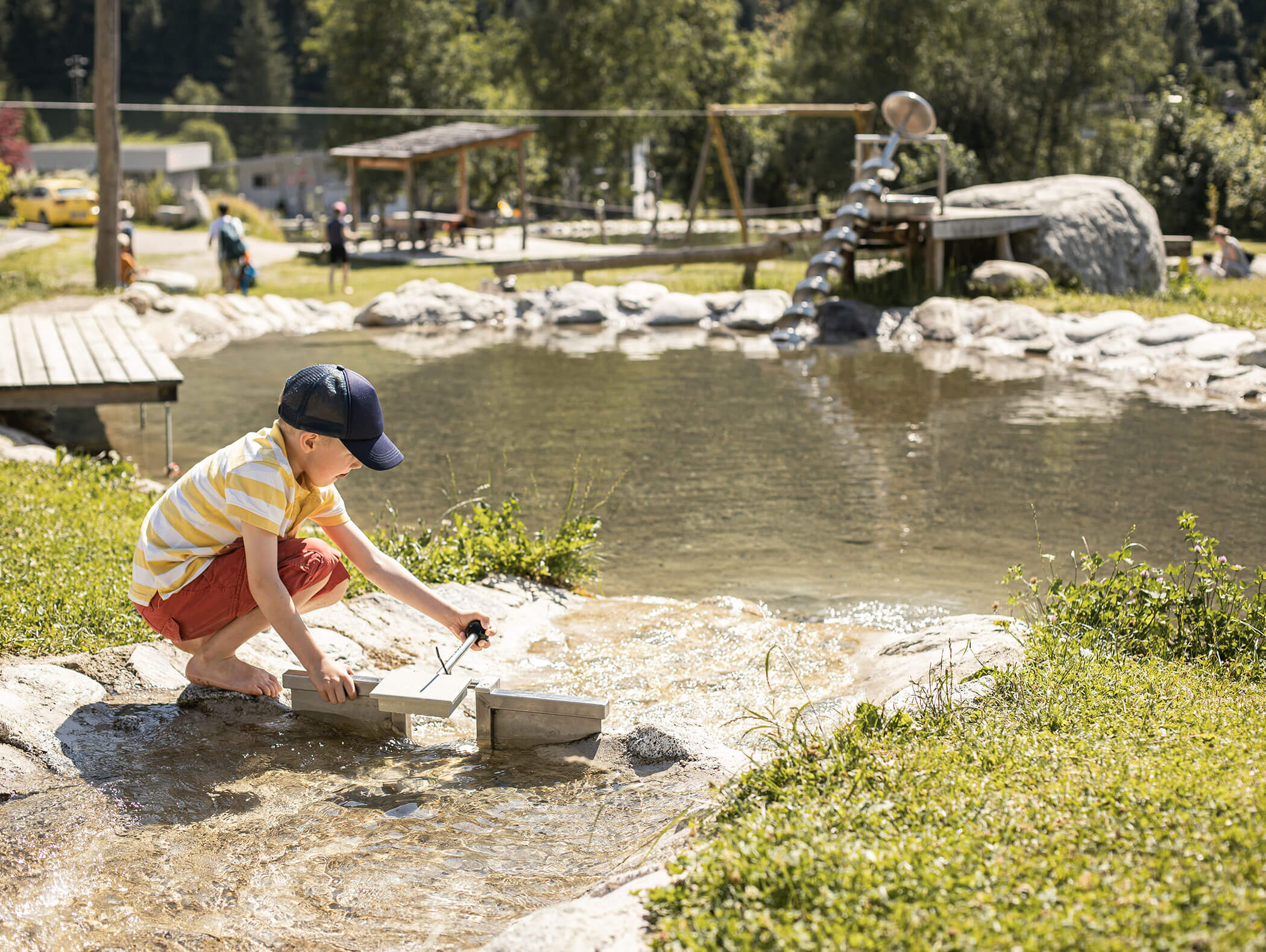 Ein Junge im gestreiften Hemd spielt an einem sonnigen Tag mit Wasser in einem Bach in der Nähe eines Teiches. - Maurlechnhof