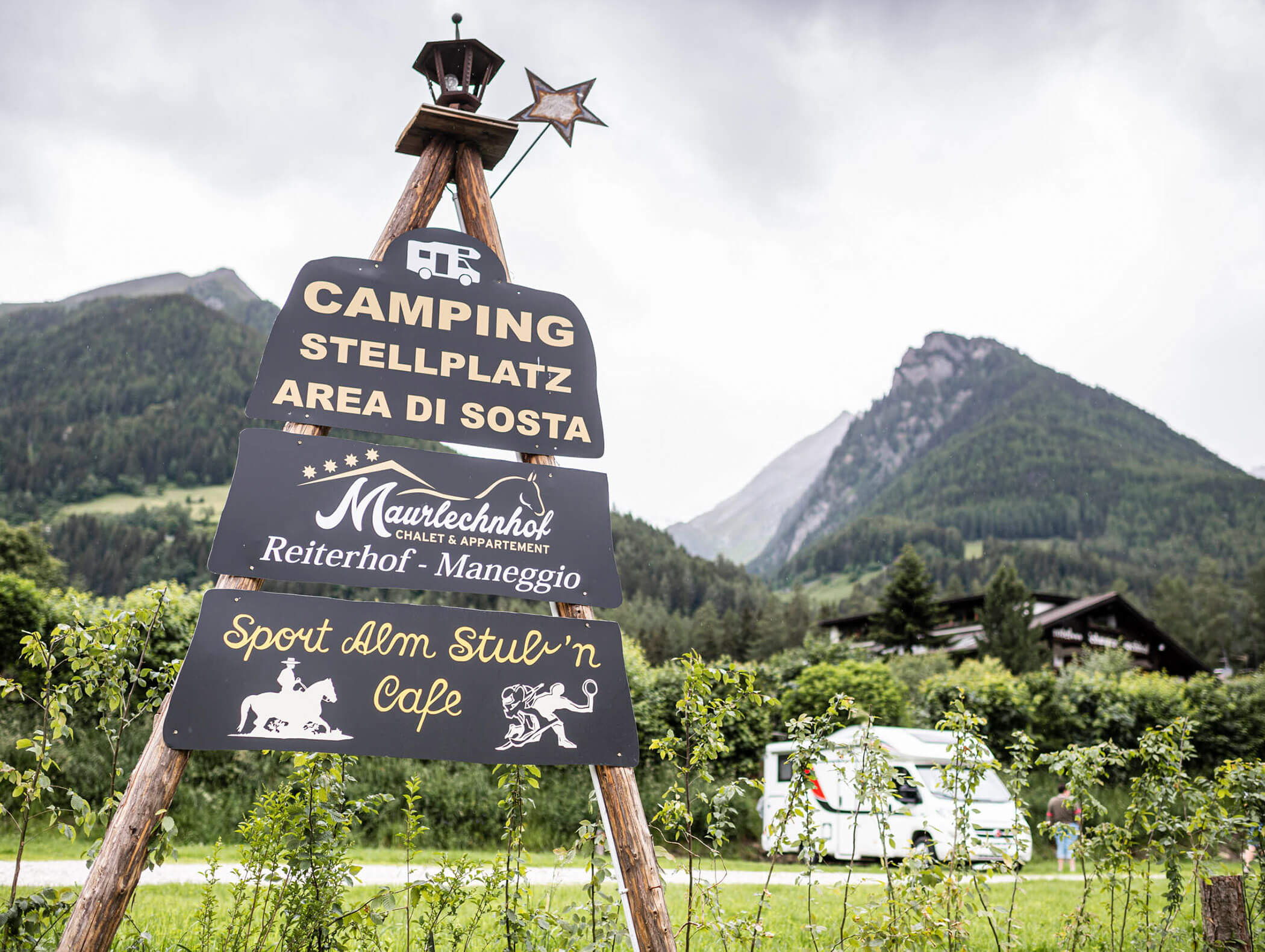 Campingplatzschild aus Holz mit mehreren Tafeln, Bergen und einem Wohnmobil im Hintergrund. - Maurlechnhof
