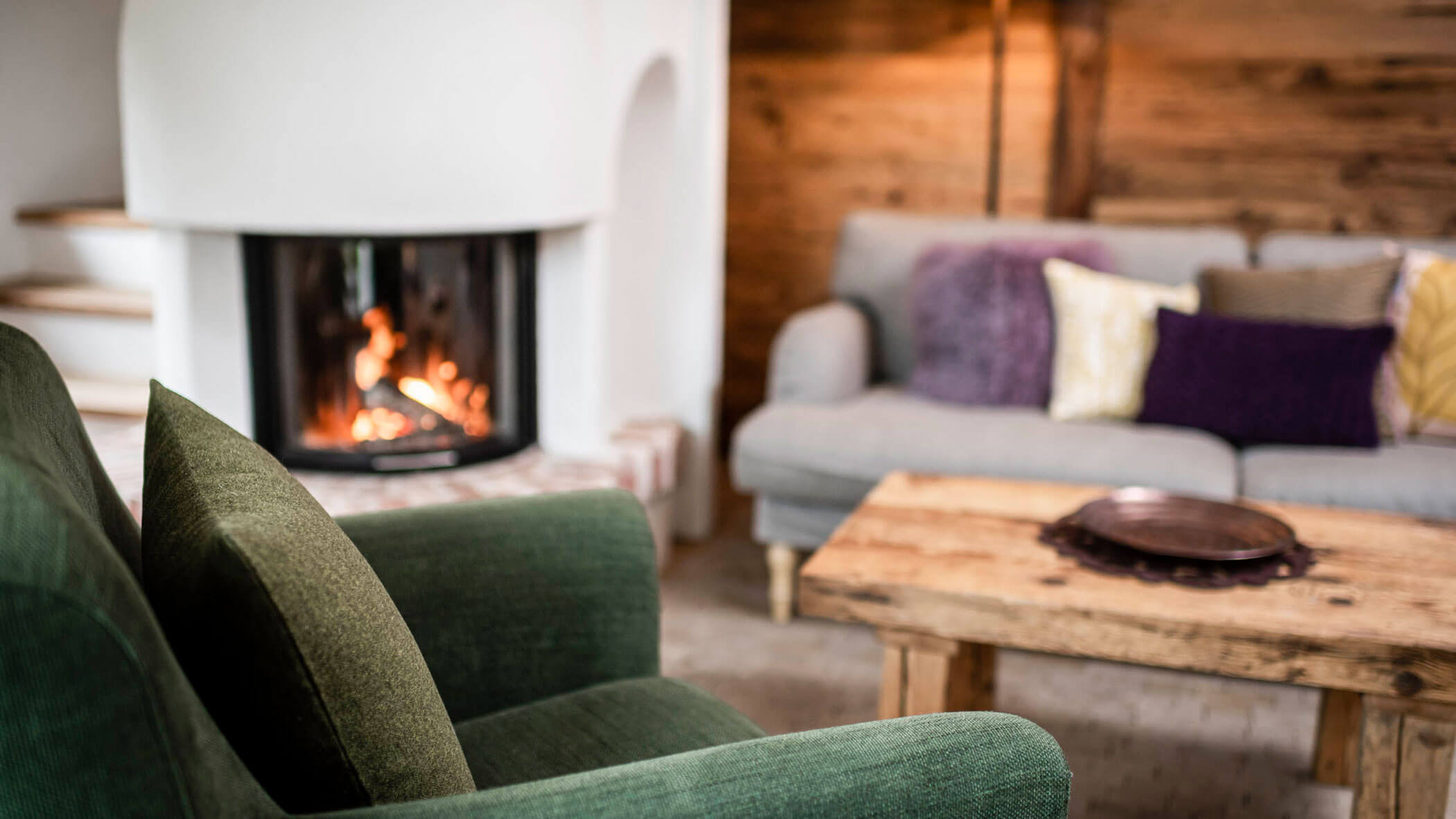 Gemütliches Wohnzimmer mit einem grünen Sessel, einem Holztisch, Sofas und einem beleuchteten Kamin im Hintergrund. - Maurlechnhof