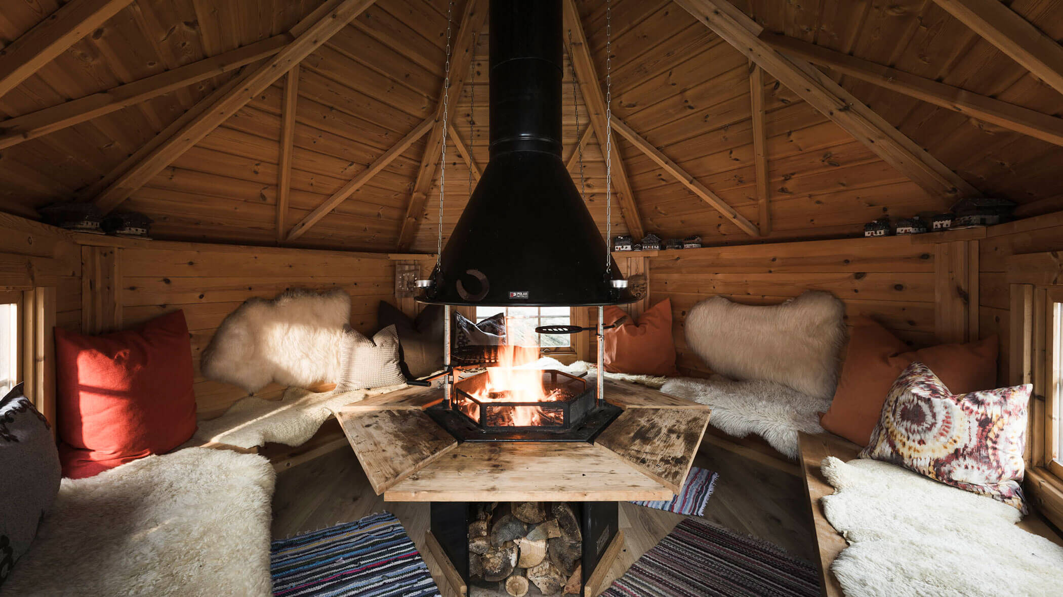 Gemütliche Holzhütte mit zentralem Kamin, Felldecken, Kissen und Bänken rund um das Feuer. - Maurlechnhof