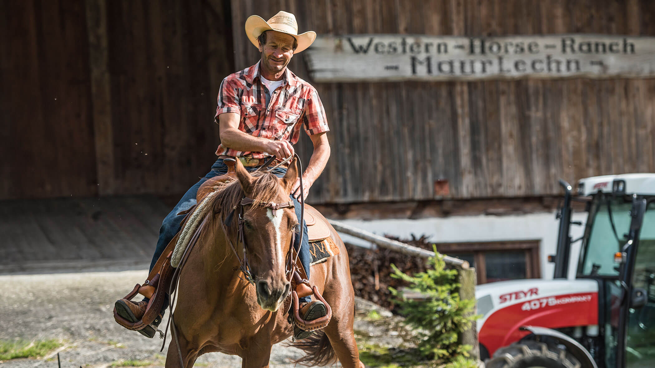 Man in a plaid shirt and cowboy hat riding a brown horse near a ranch sign and a red tractor. - Maurlechnhof