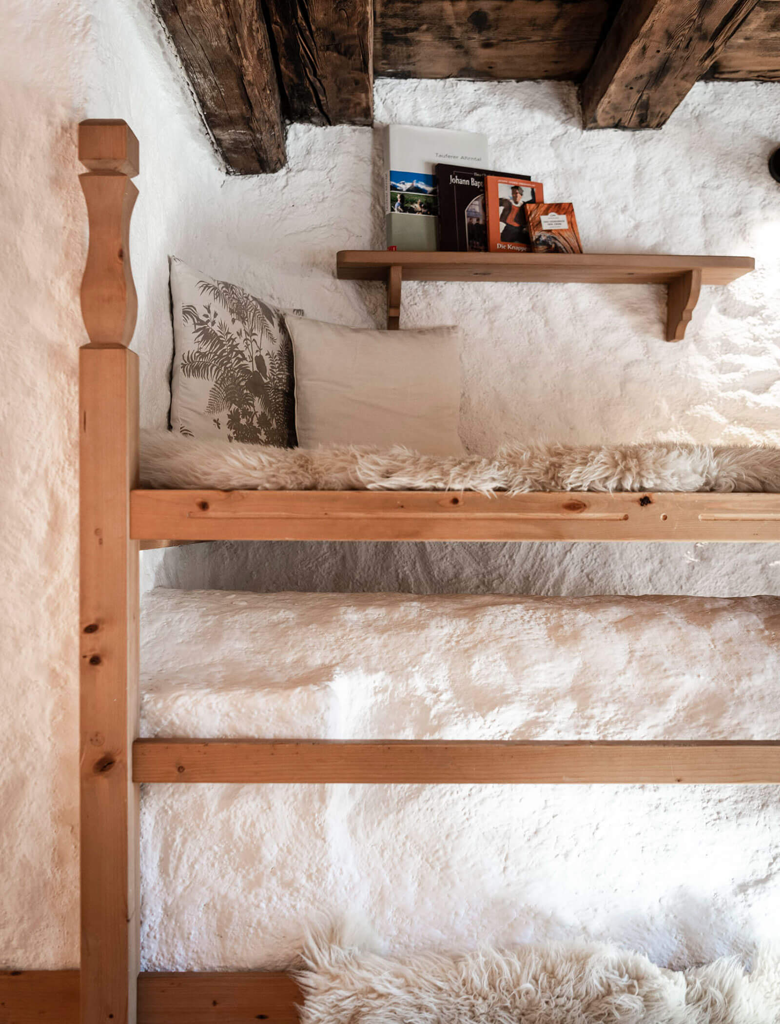 Etagenbett aus Holz mit flauschiger weißer Bettwäsche, rustikaler weißer Wand und einem kleinen Regal mit Büchern darüber. - Maurlechnhof