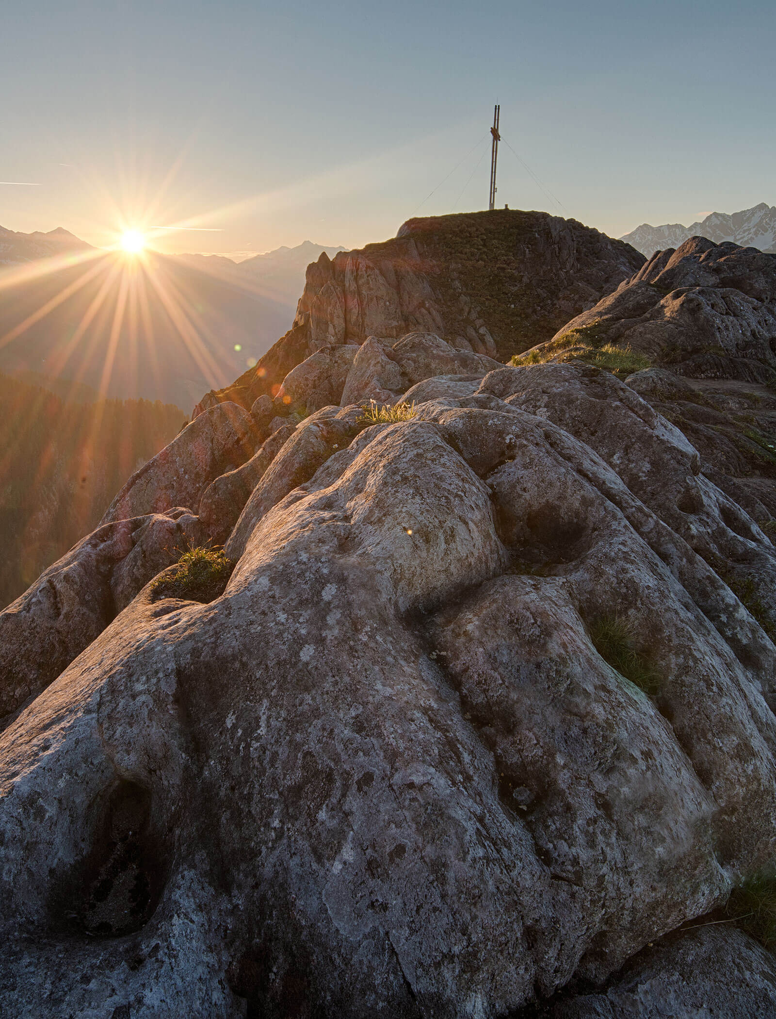 Sonnenaufgang über einem felsigen Berggipfel mit einem Kreuz, das lange Schatten wirft und die Landschaft beleuchtet. - Maurlechnhof