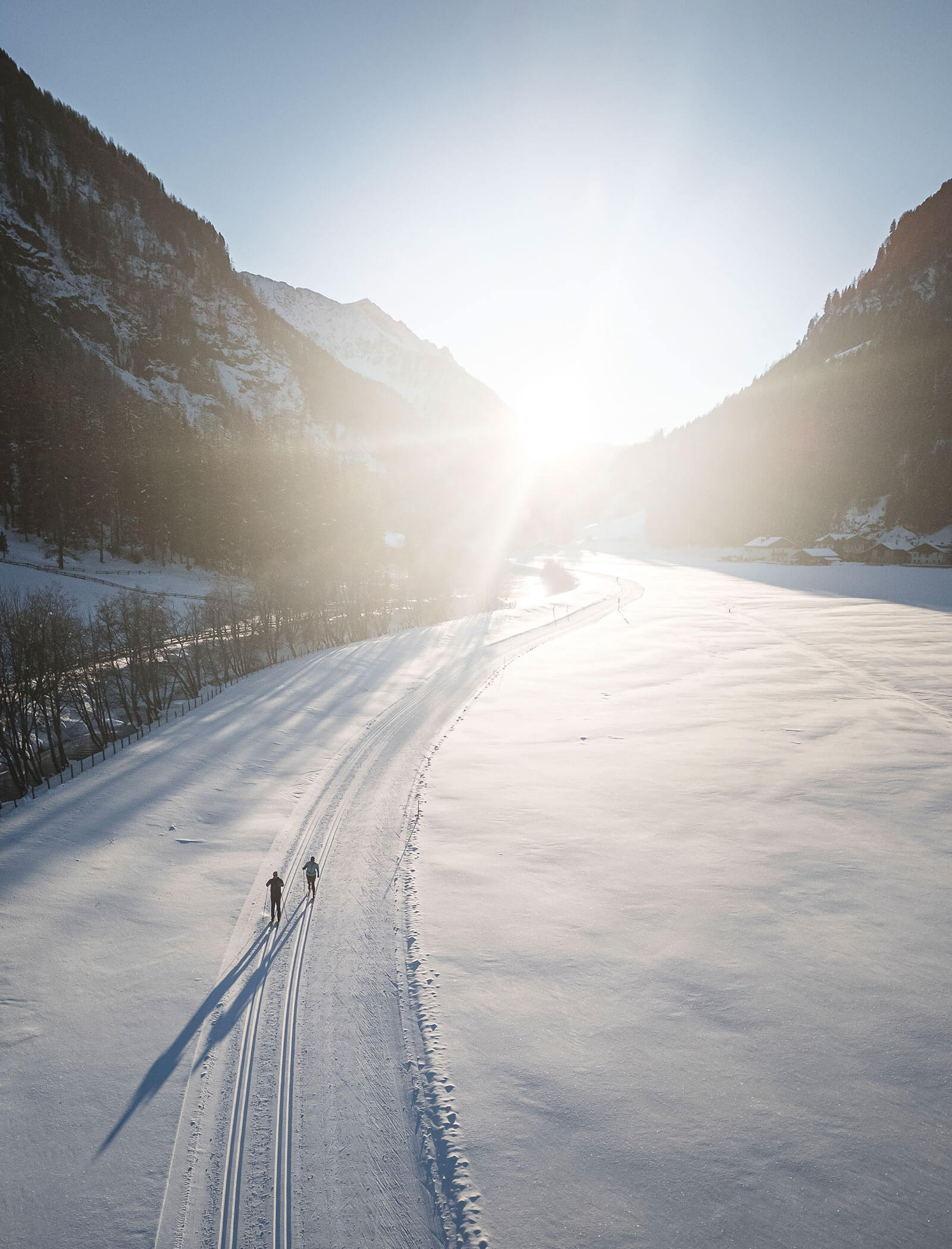 Zwei Langläufer auf einer verschneiten Loipe durch ein Tal bei Sonnenaufgang, umgeben von Bergen. - Maurlechnhof